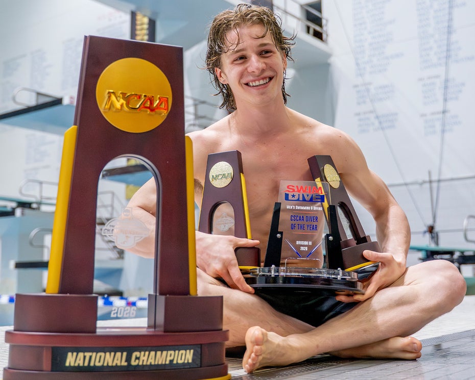 A young man in a swimsuit sits on a pool deck, smiling and holding several NCAA diving trophies, with a large “National Champion” trophy in the foreground and a diving board in the background.
