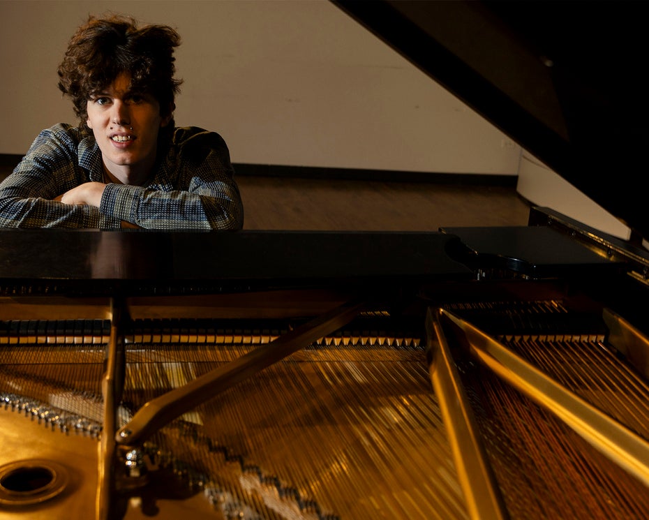 A person with curly hair leans on a grand piano, looking toward the camera. The pianos strings and internal components are visible in the foreground.