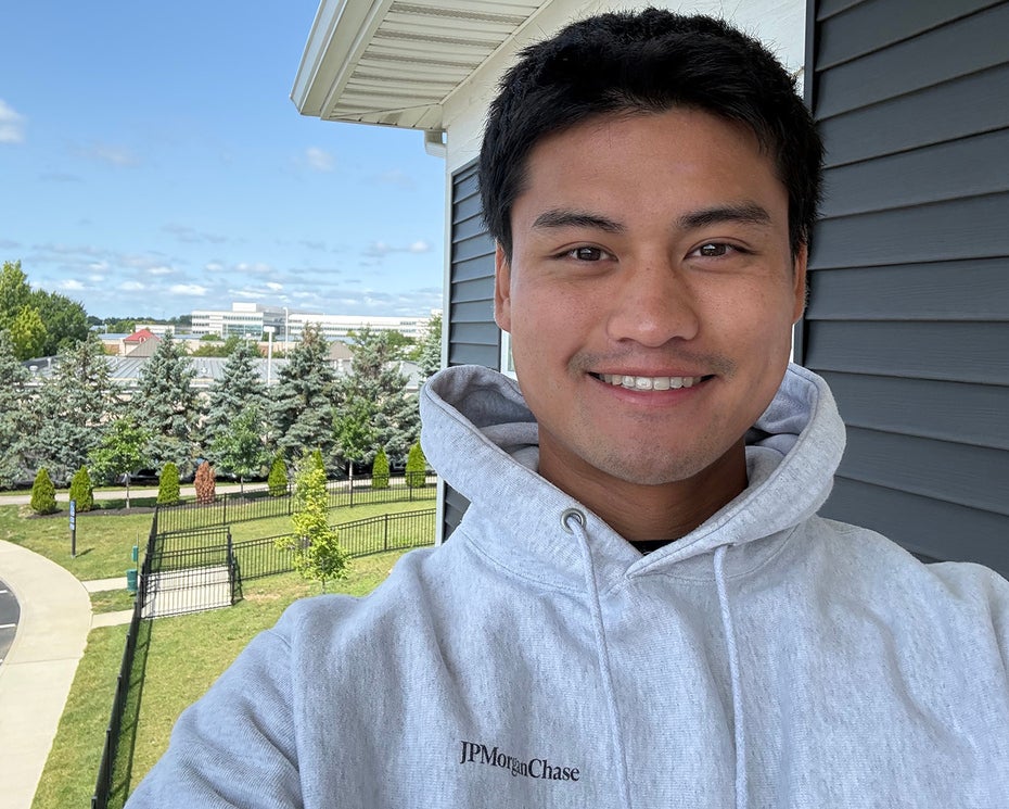 A person wearing a light gray hoodie smiles for a selfie outside near a building, with trees, grass, and a parking lot in the background under a blue sky.