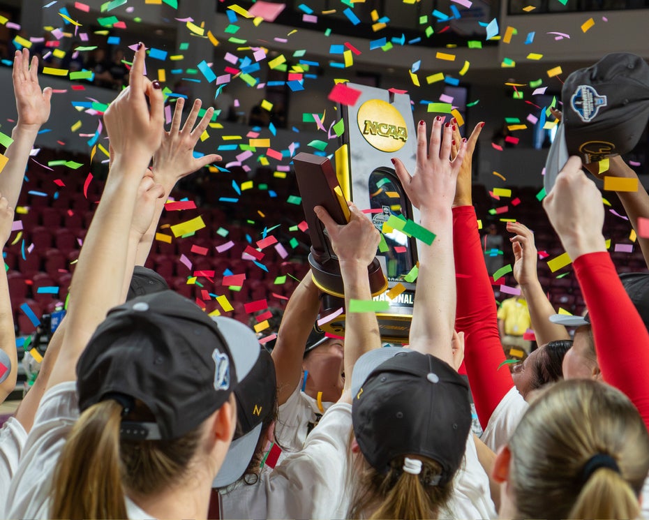 A sports team wearing hats raises a trophy together amid falling confetti in an indoor arena, celebrating a championship victory.