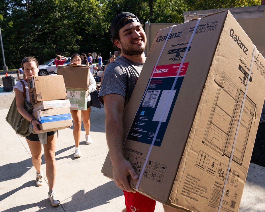 People carry large boxes and belongings outdoors on a sunny day, possibly moving into a dorm or apartment.