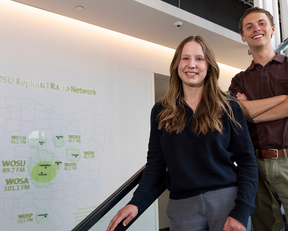 Two people stand side by side on a staircase indoors, smiling at the camera. A diagram is visible on the white wall behind them.