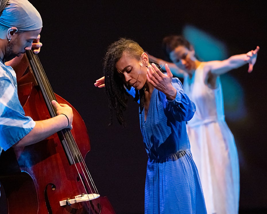 A musician plays an upright bass while two dancers, one in blue and one in white, perform expressive movements on a dimly lit stage.