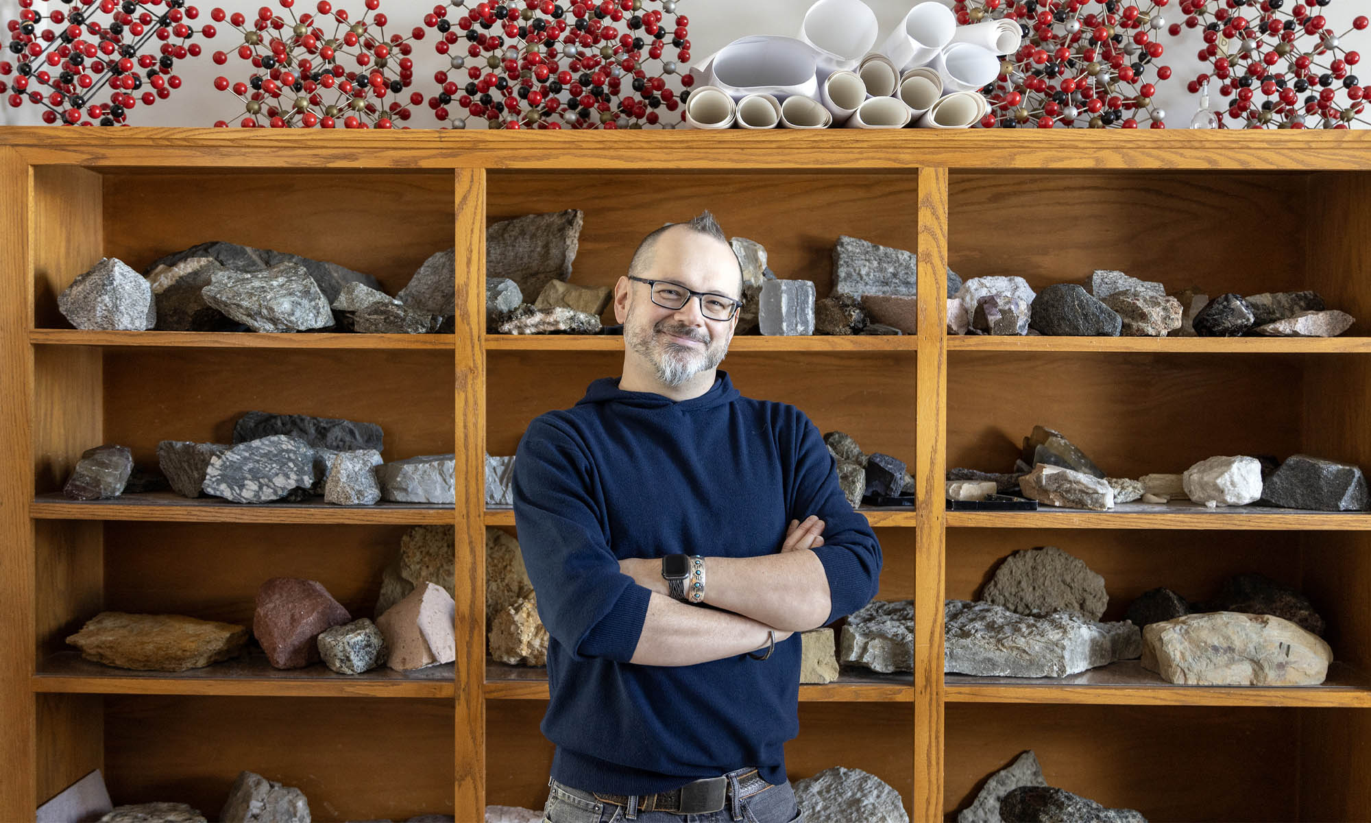 Photo of Geosciences Associate Professor Erik Klemetti standing in front of several wood shelves displaying various types of rocks
