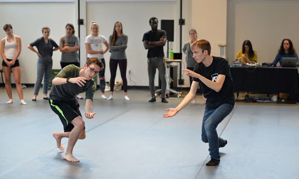 Student practice choreography in a rehearsal space while their classmates observe.