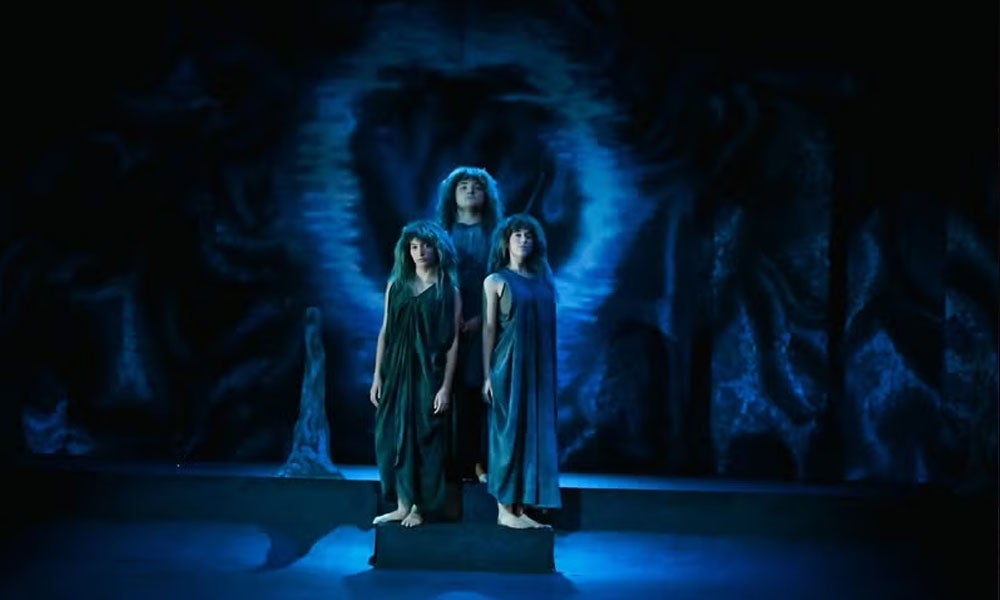 Three actors in gray cloaks stand in blue light on a stage designed to look like a cave.