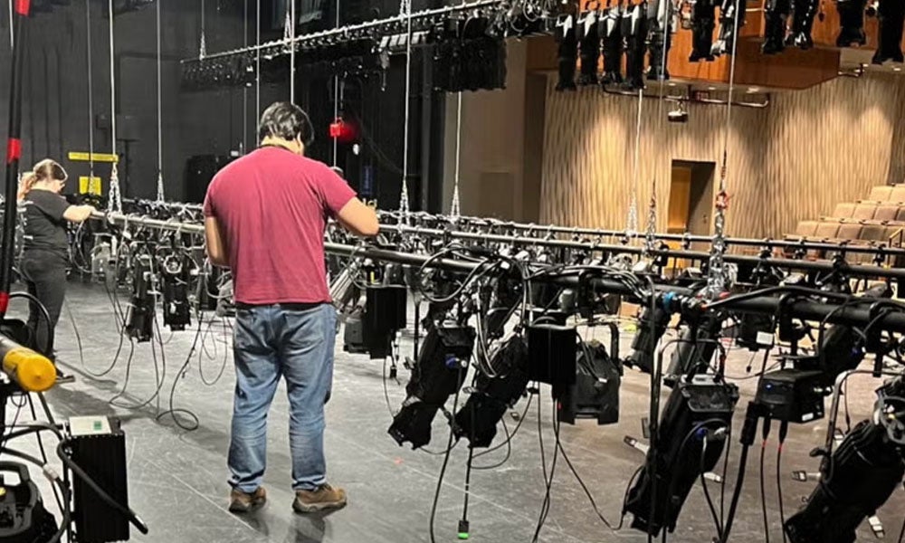 A man stands on a theater stage surrounded by lights and rigging.