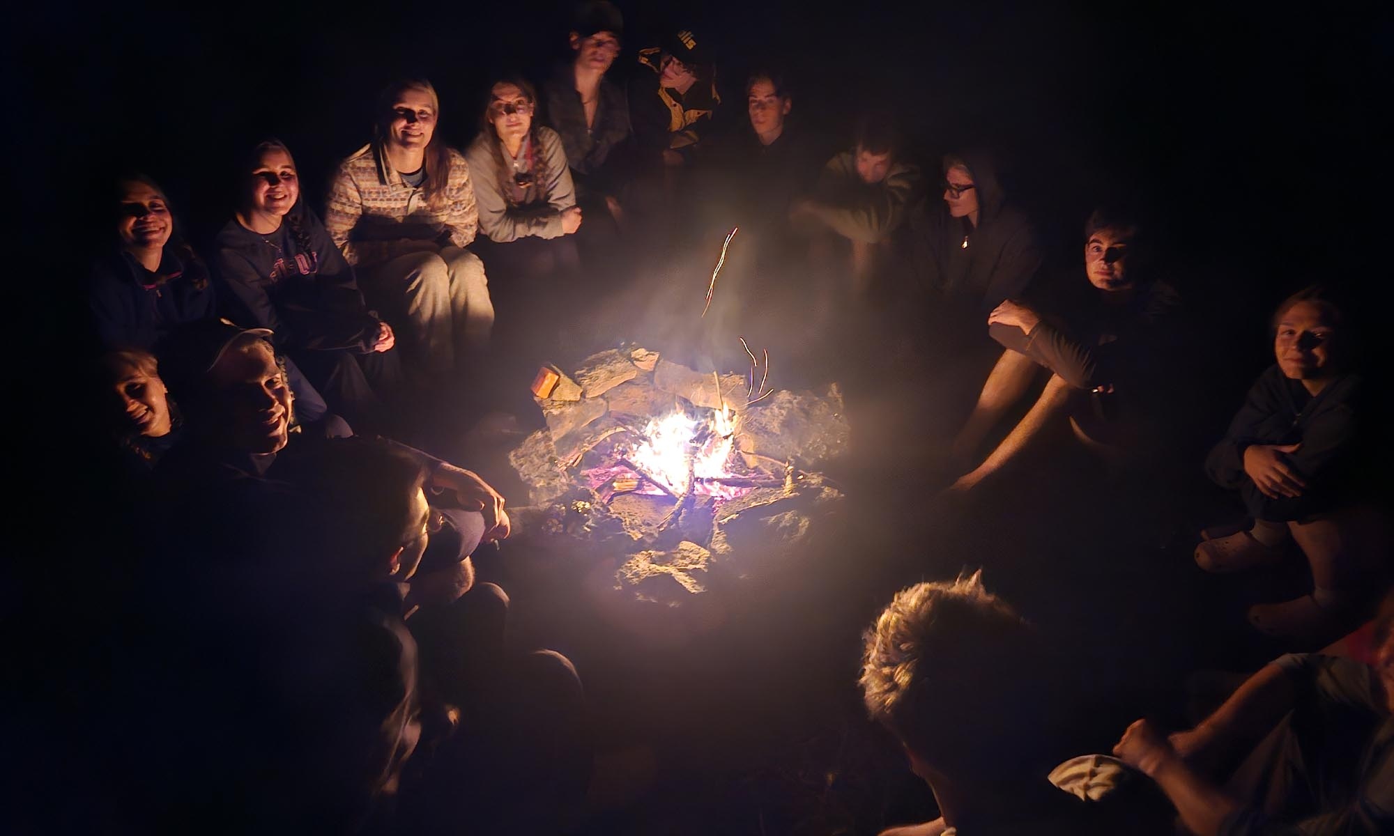 A group of friends smiles while sitting around a camp fire.