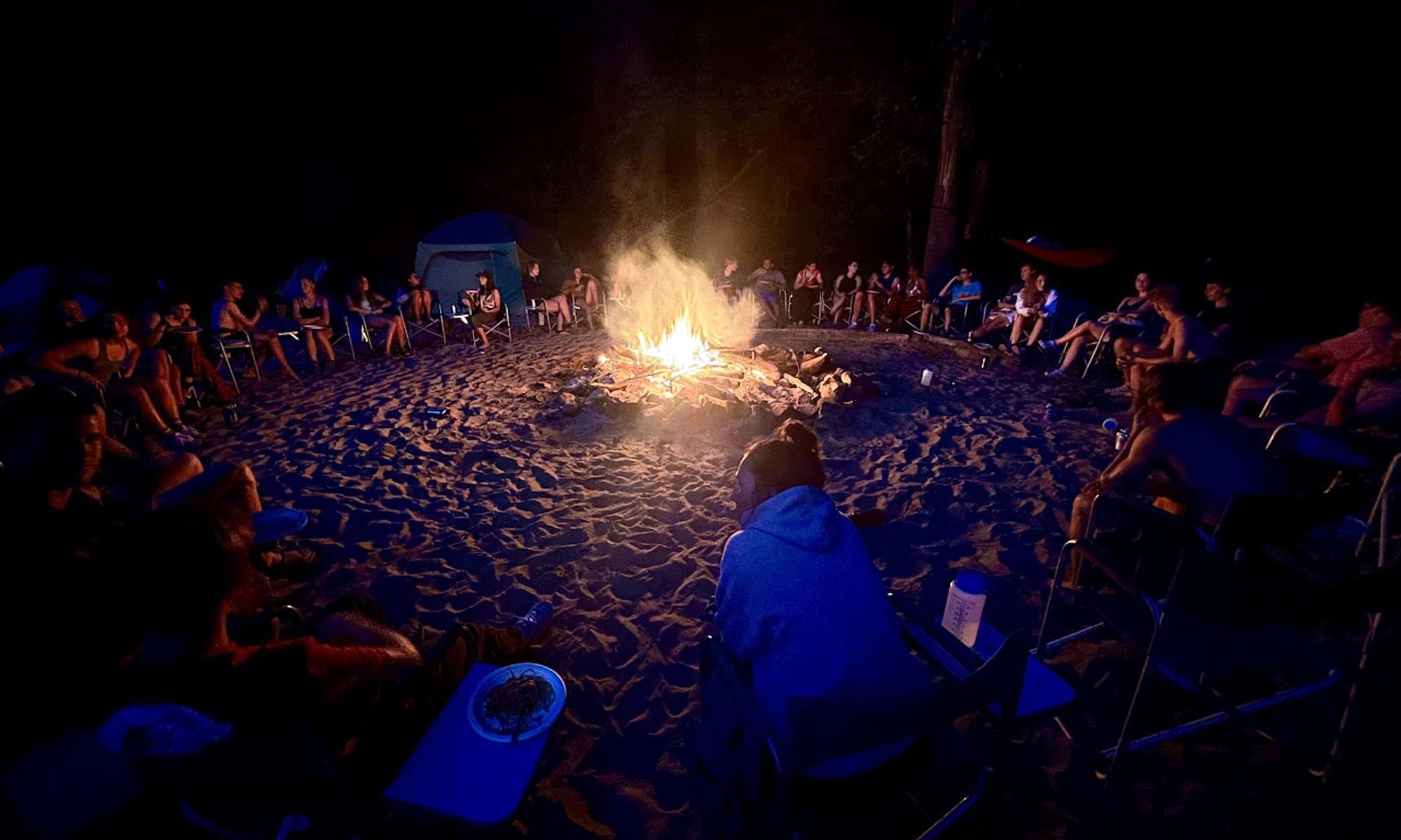 A large group sits around a bonfire at night after a day of rafting.