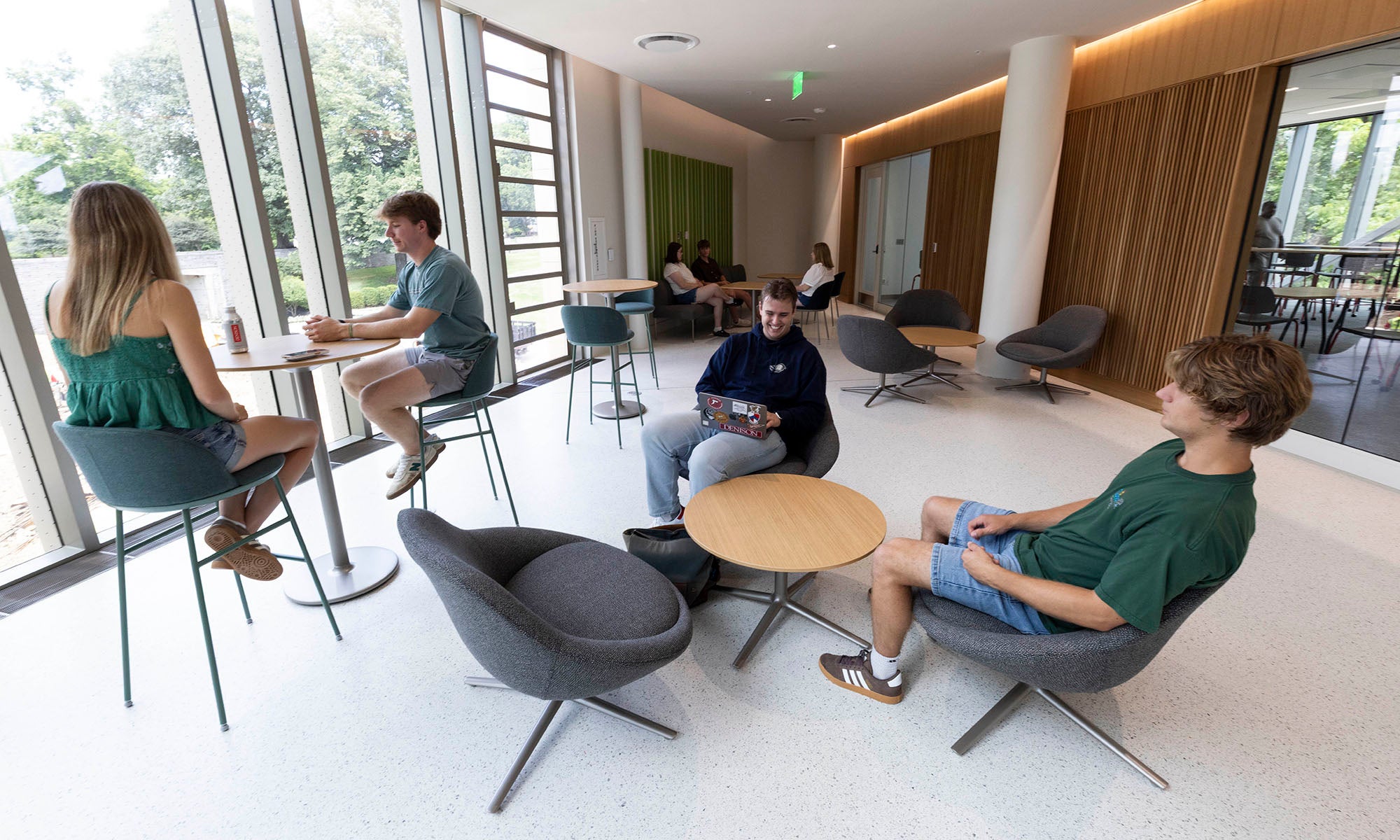 Four people sit in a modern lounge area with large windows, talking and using devices. Other people are seen in the background.