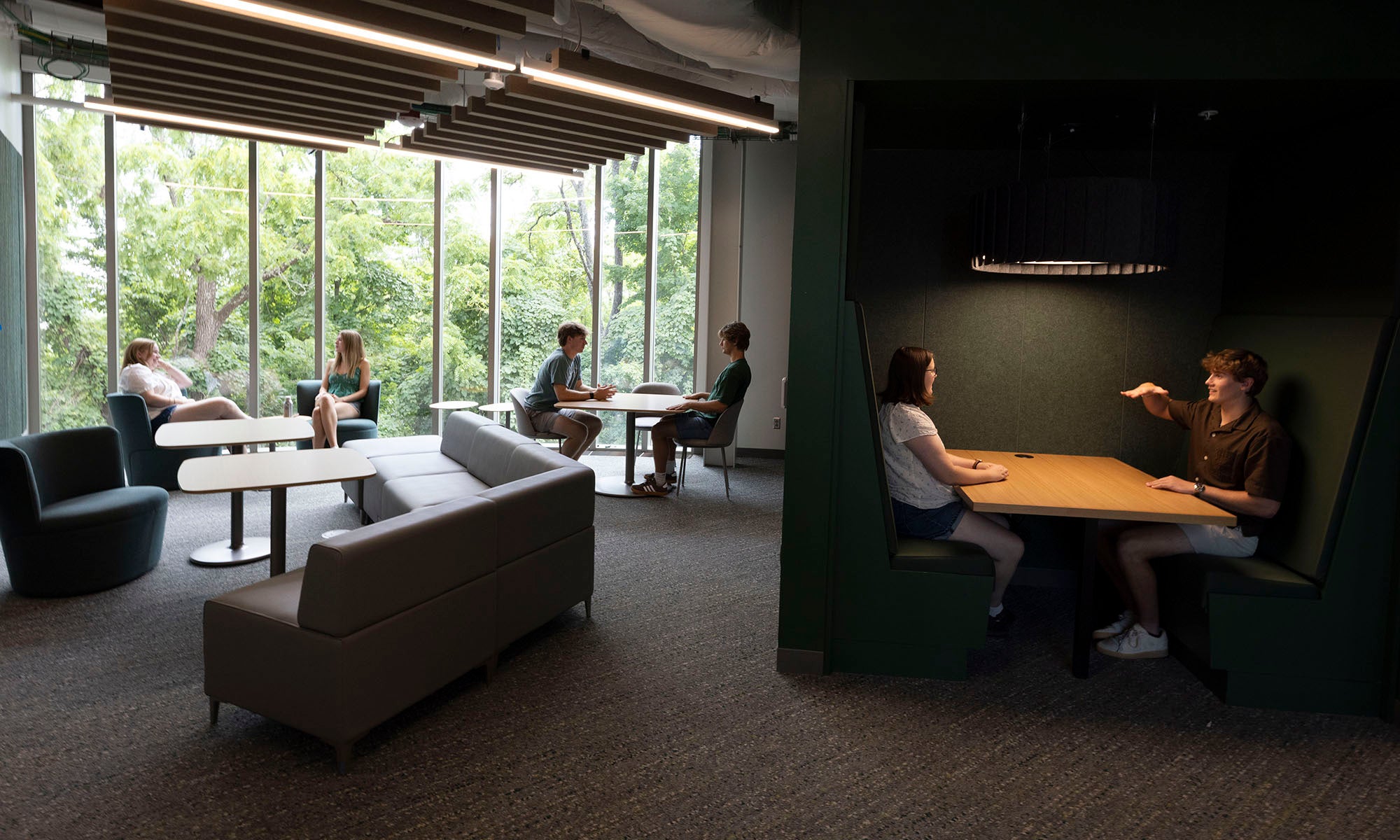 People sit in pairs at tables and booths in a modern lounge area with large windows, natural light, and greenery visible outside.