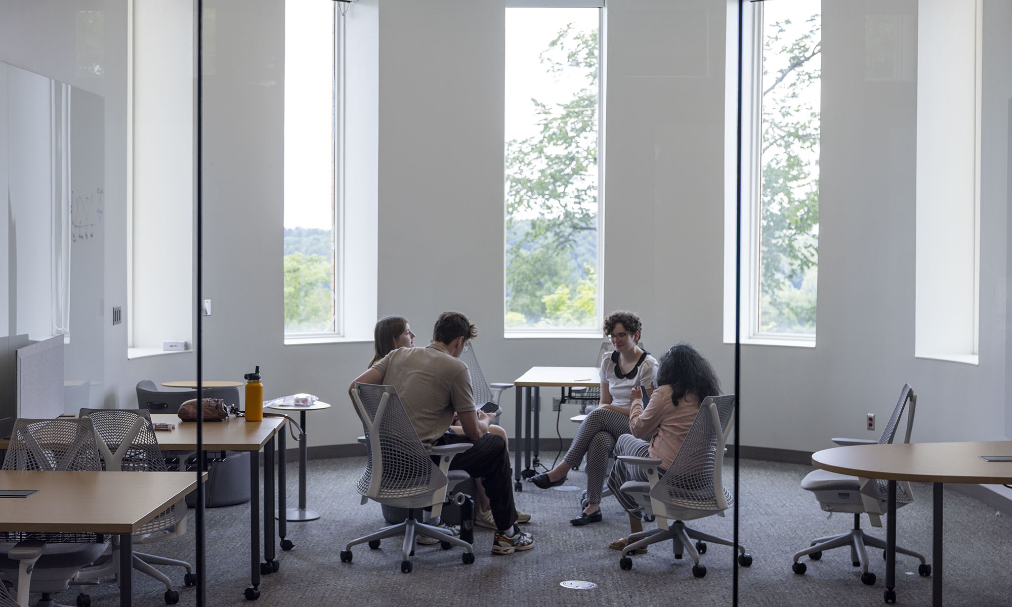 Four people sit in a circle having a discussion in a modern, glass-walled office with large windows and empty desks around them.