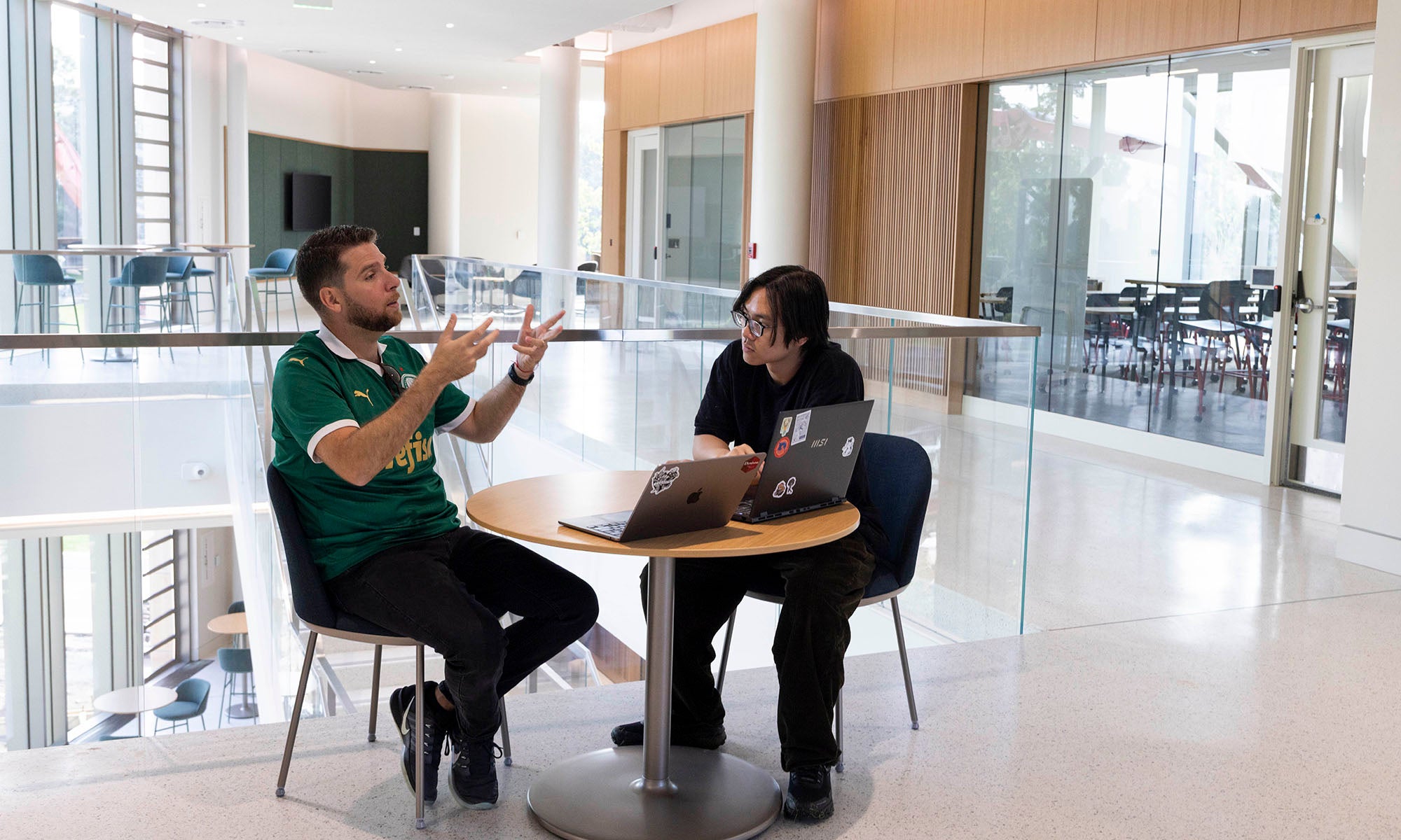 Two people sit at a round table with laptops, engaged in discussion in a modern, bright indoor space with large windows.