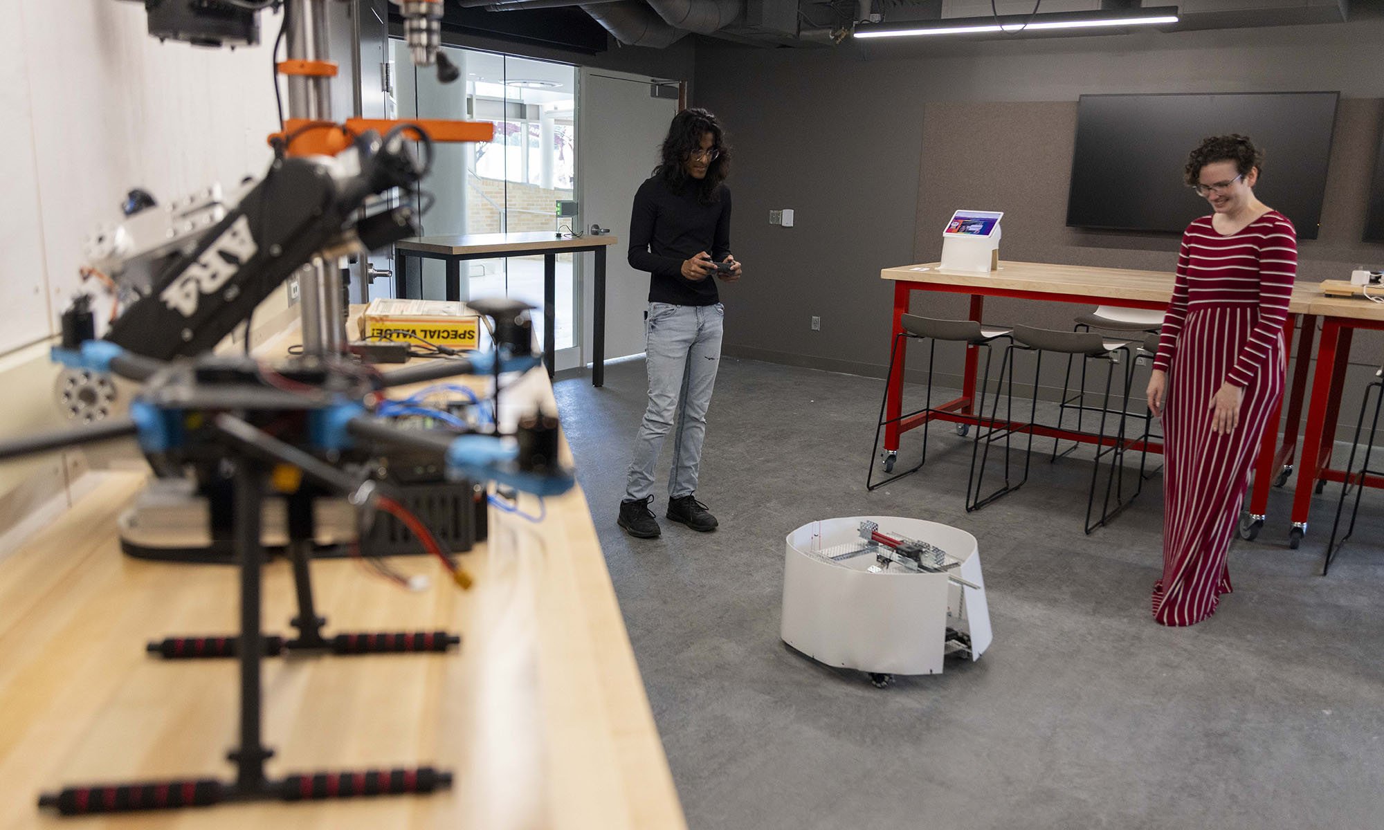 Two people observe a round robot in a modern workspace, with a drone and equipment visible on a counter in the foreground.