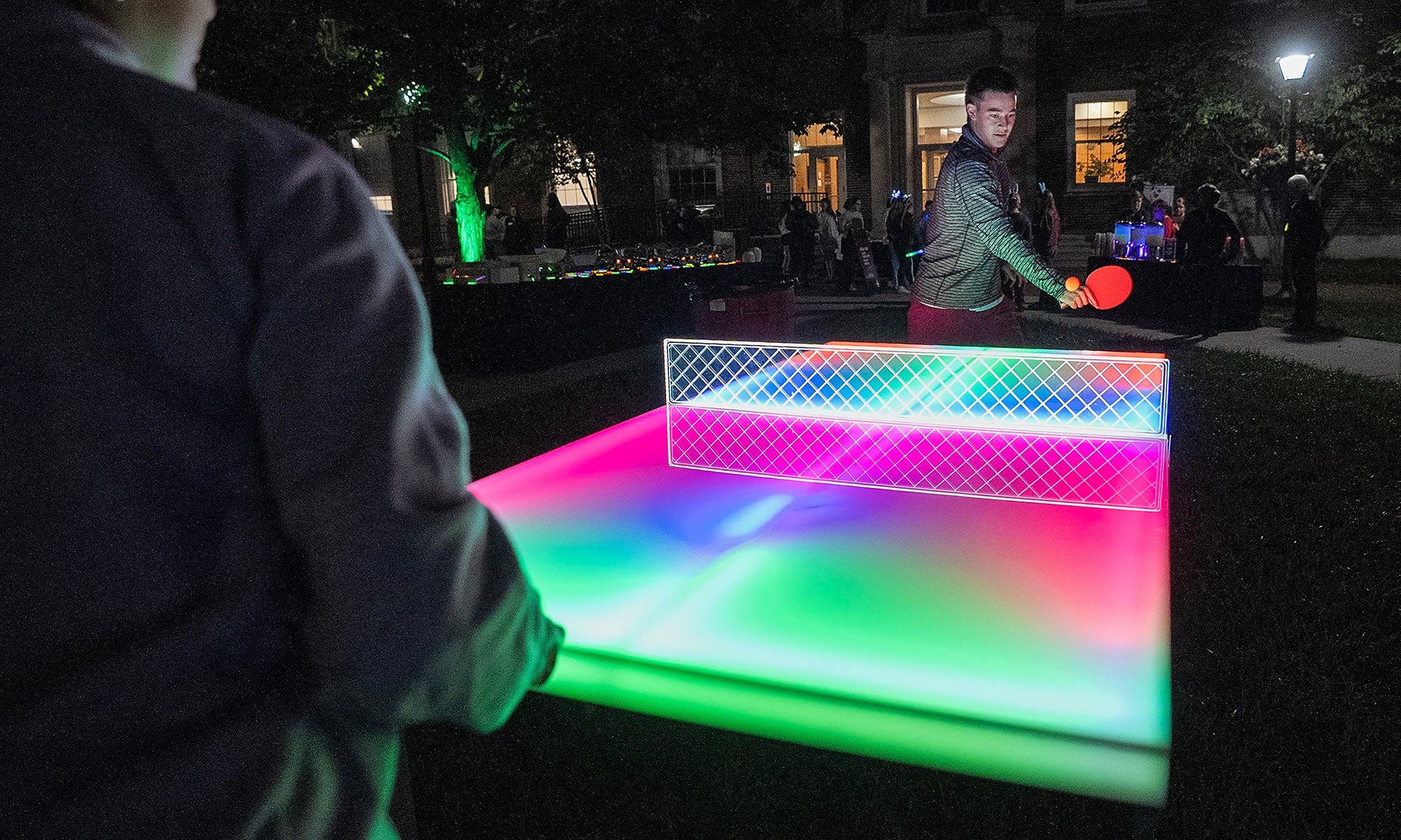 Two people play ping-pong at night on a glowing, brightly colored ping pong table.