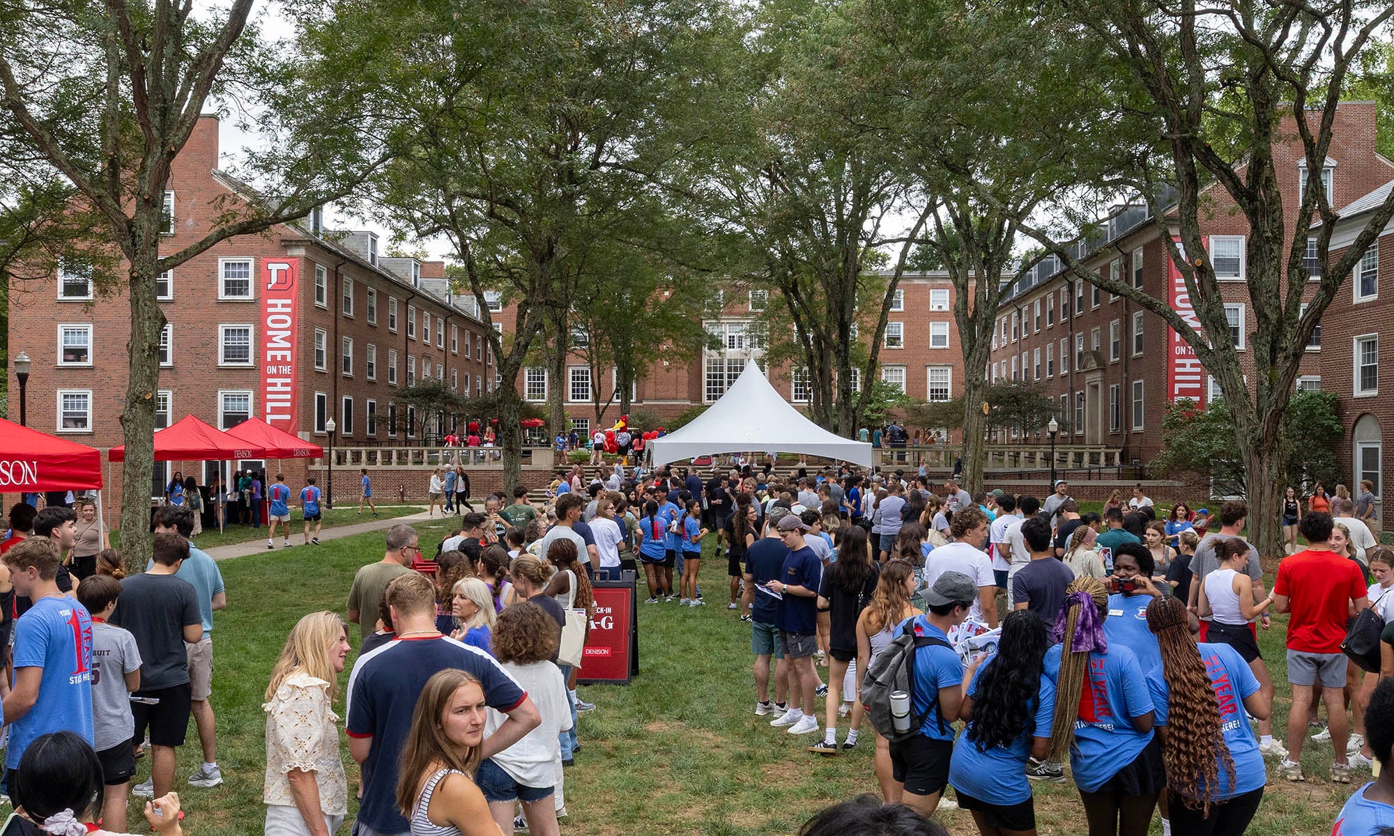 A large group of people stands in lines on a grassy quad between brick dorm buildings, attending campus programs under a white tent and red canopies. Trees provide shade overhead.