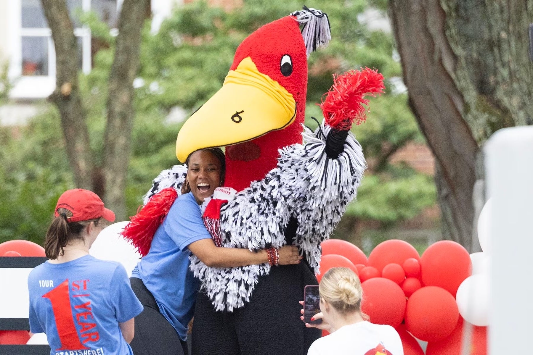 A student smiles and hugs Buzzy while surrounded by balloons on the quad