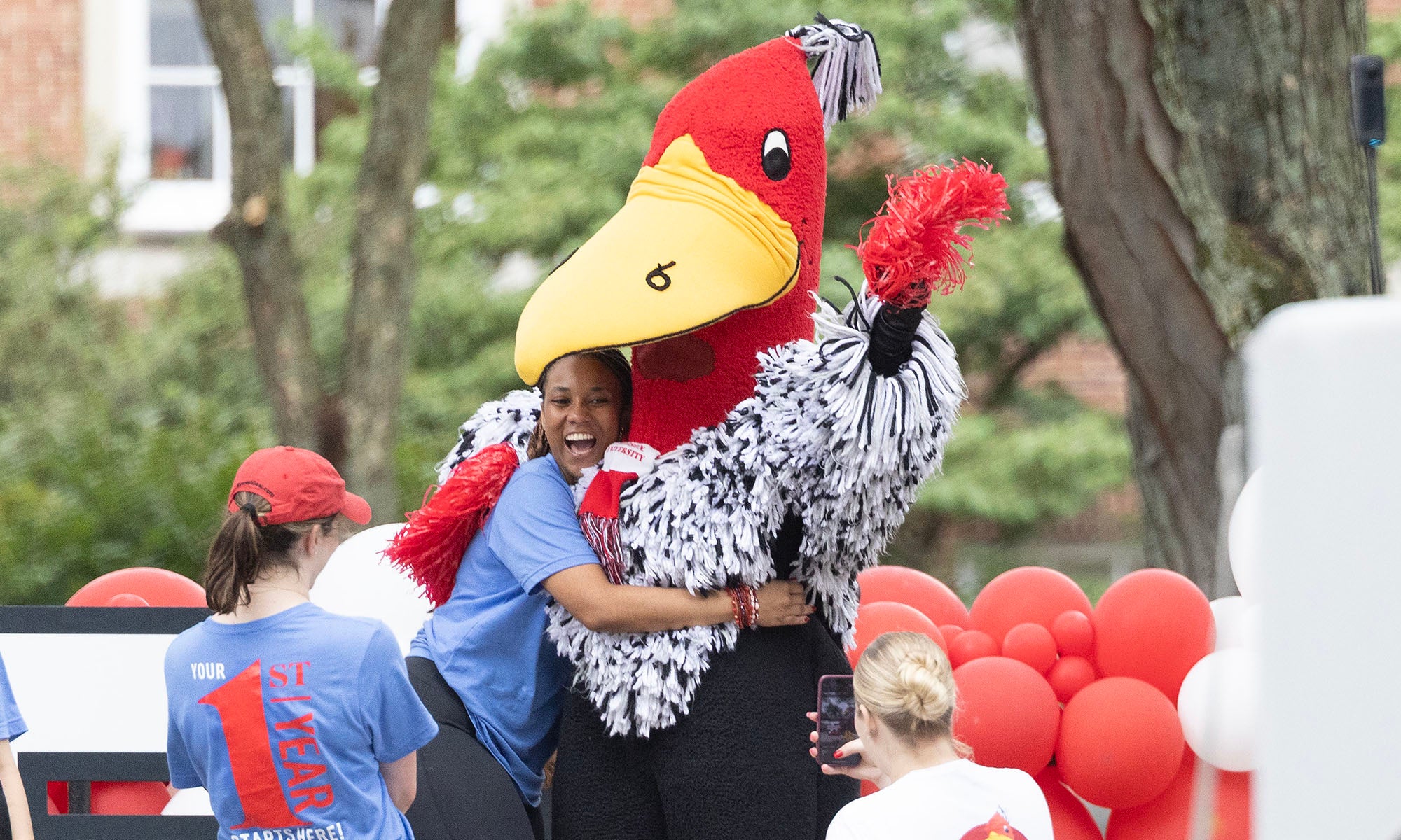 A person in a large red bird mascot costume hugs a smiling woman outdoors, surrounded by people wearing blue and red shirts, with red and white balloons—a joyful scene that highlights how campus programs help build connections at Denison.
