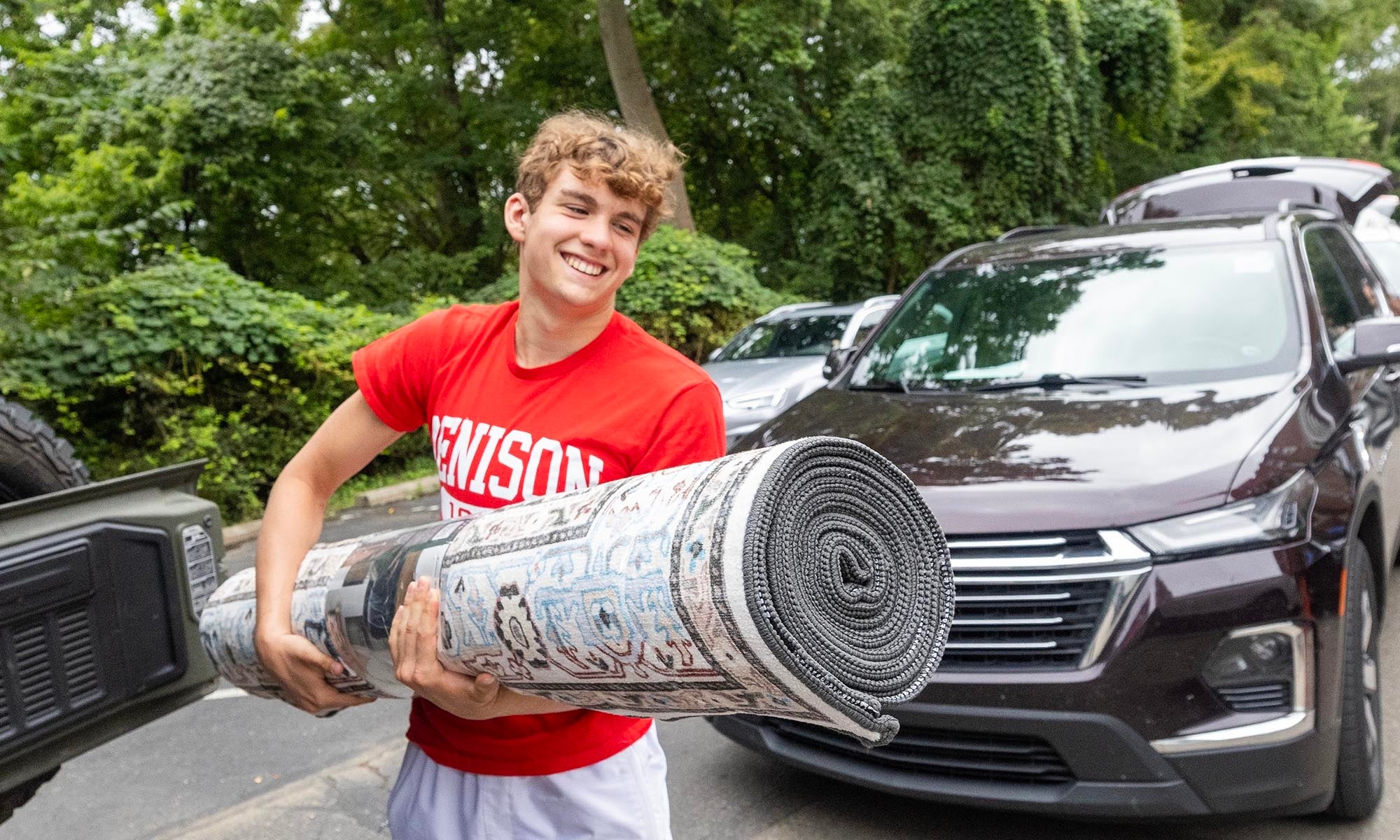 A young man in a red shirt carries a large rolled-up floor rug during campus move-in.