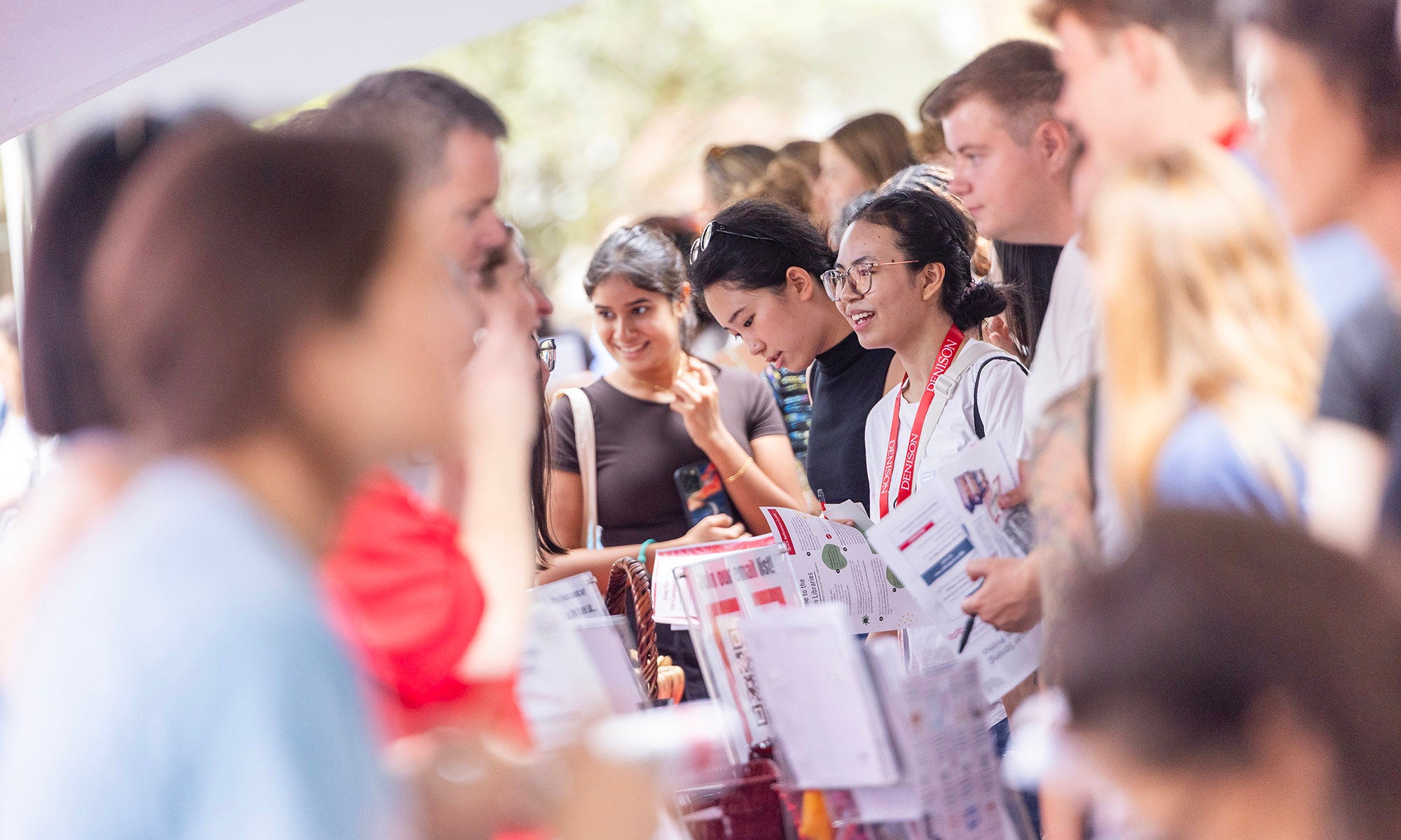 A group of people gather around tables with informational brochures and papers, engaging in conversation at a busy event focused on campus programs and building student connections.