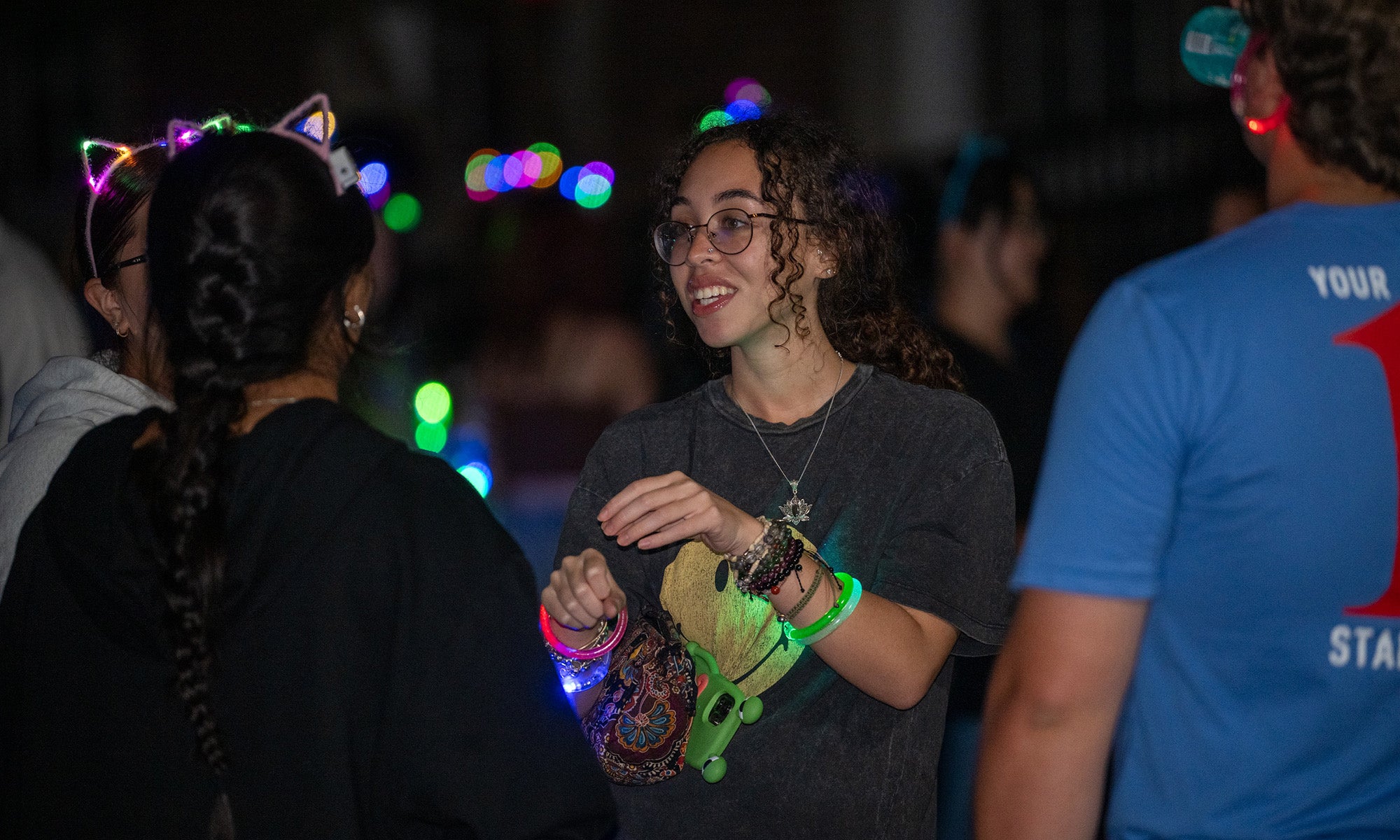 A group of people stands together at night. One person in the center, wearing glasses, a T-shirt, and glow bracelets, is talking to someone with cat ear headbands at a lively campus program with colorful lights in the background.