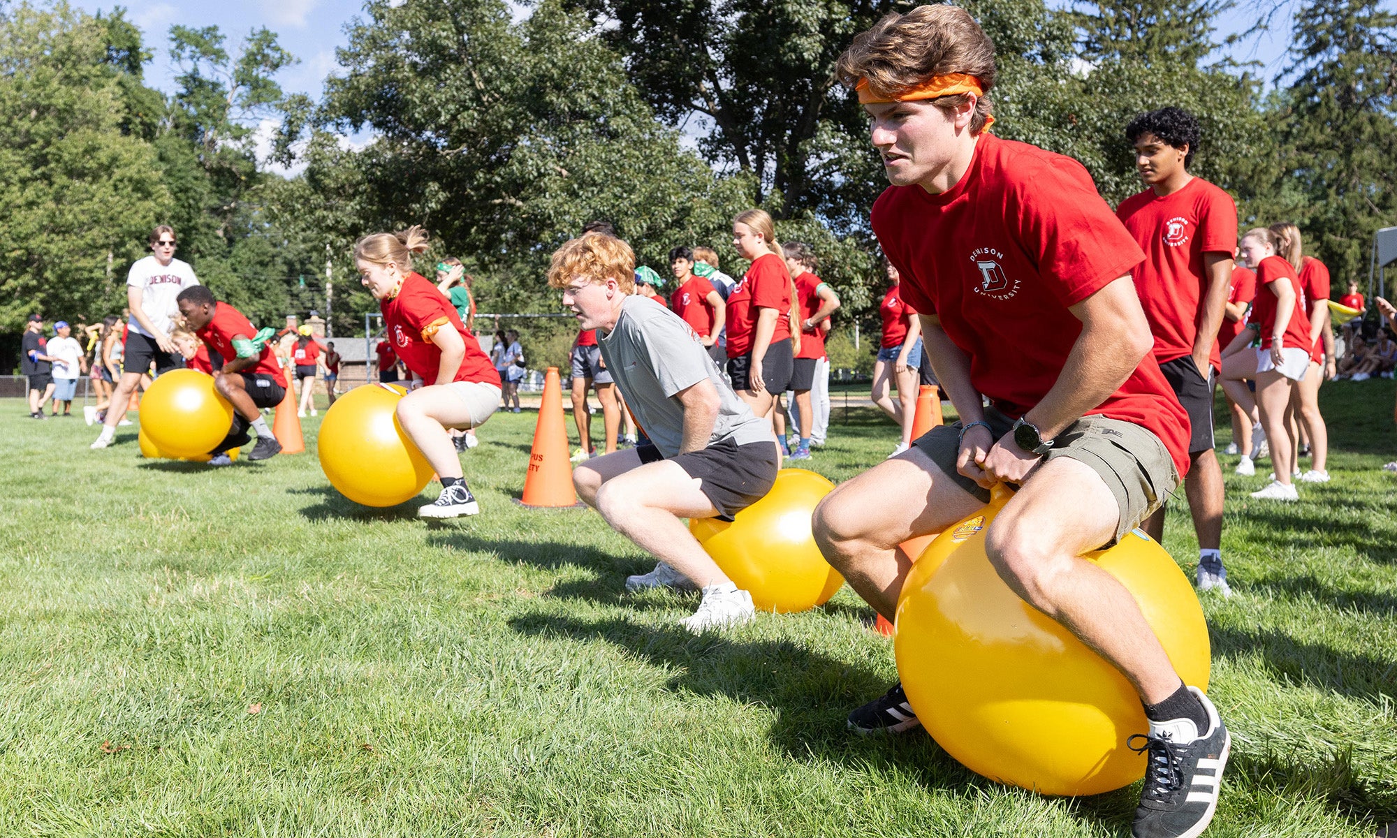 A group of young people outdoors, likely part of campus programs, participate in a race by bouncing on large yellow exercise balls as others watch and cheer on the grassy field.