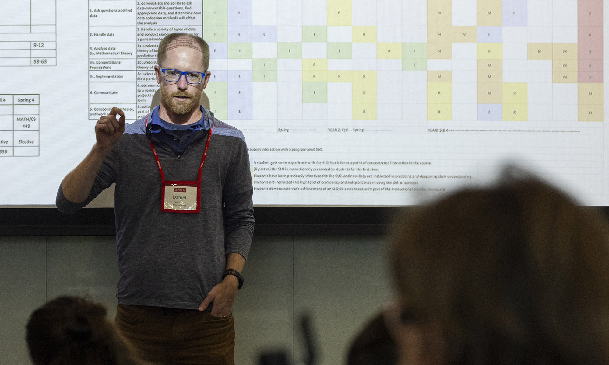 A man with glasses and a name tag gives a presentation in front of a projected spreadsheet.