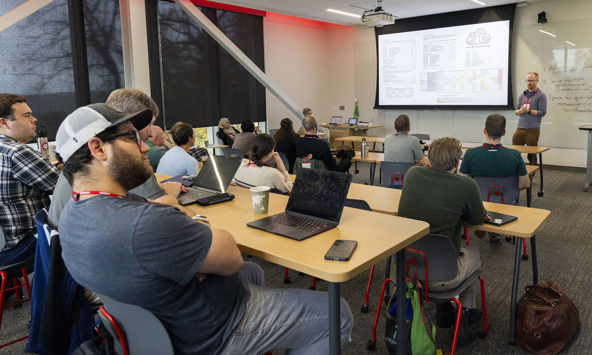A speaker presents to a small audience in a classroom with laptops open on desks.