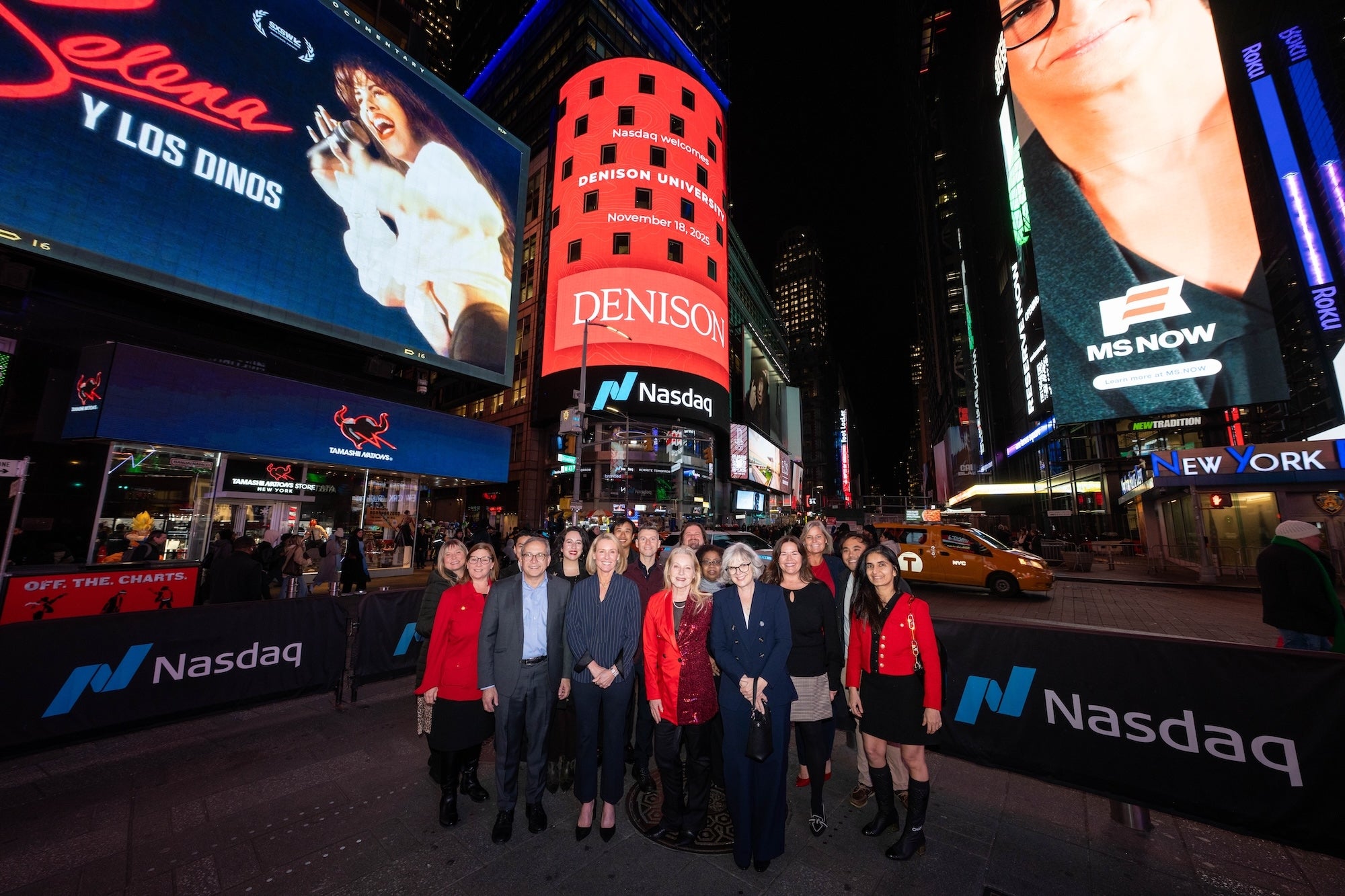 A group of people stands in Times Square at night in front of Nasdaq signs, surrounded by illuminated billboards and advertisements, including Denison.