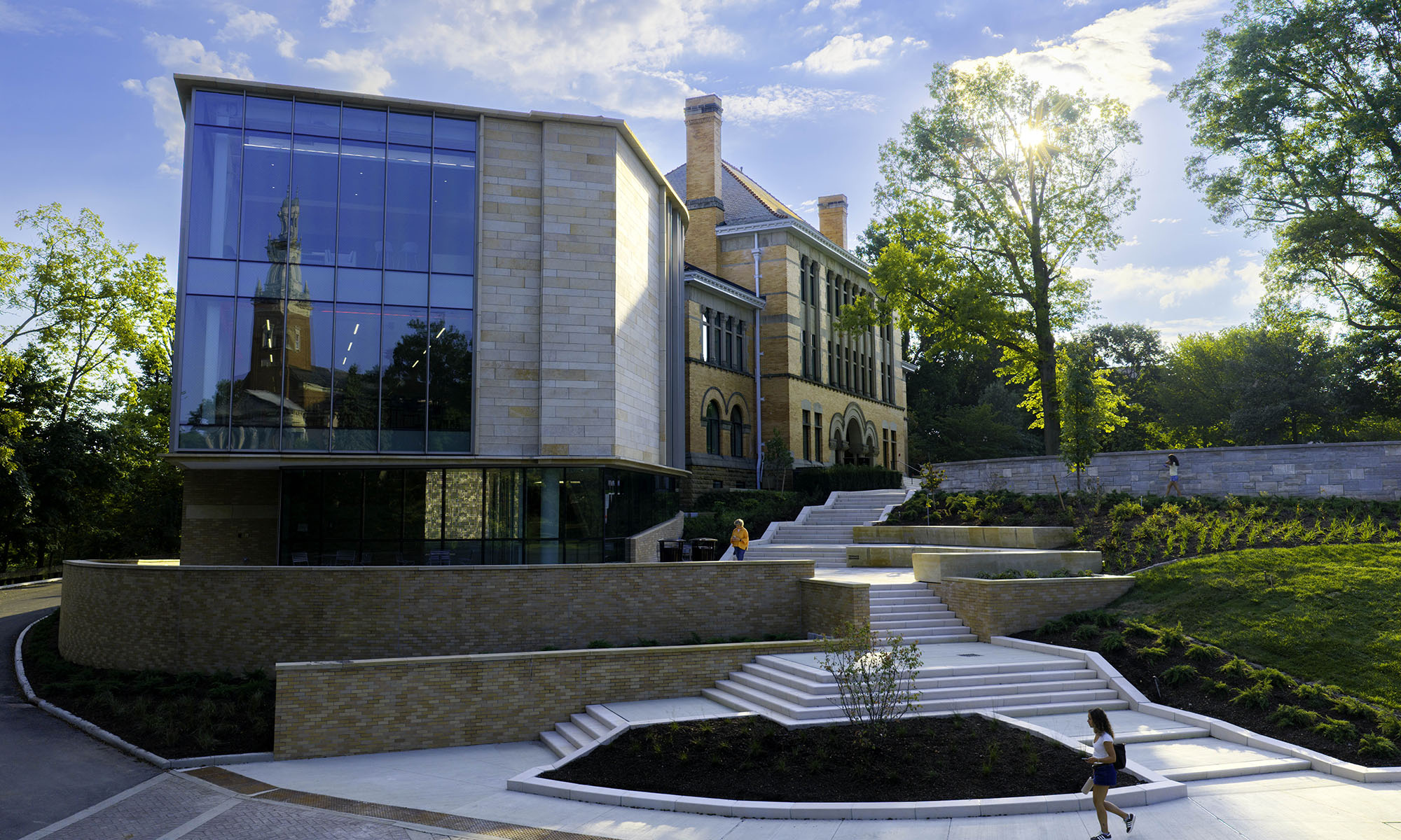 Modern building with large glass windows and brick sections, surrounded by landscaped stairs, greenery, and a person walking in the lower right corner under a sunny sky.