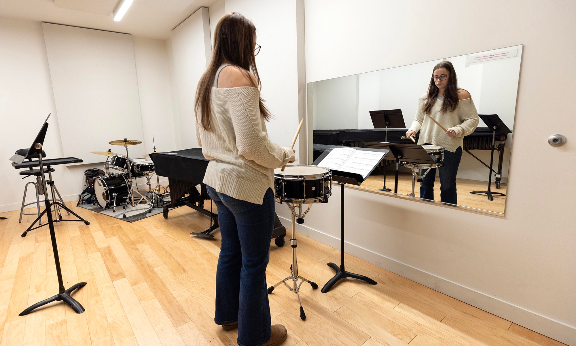 A student practices snare drum in a music practice room, standing in front of a mirror with sheet music on a stand.