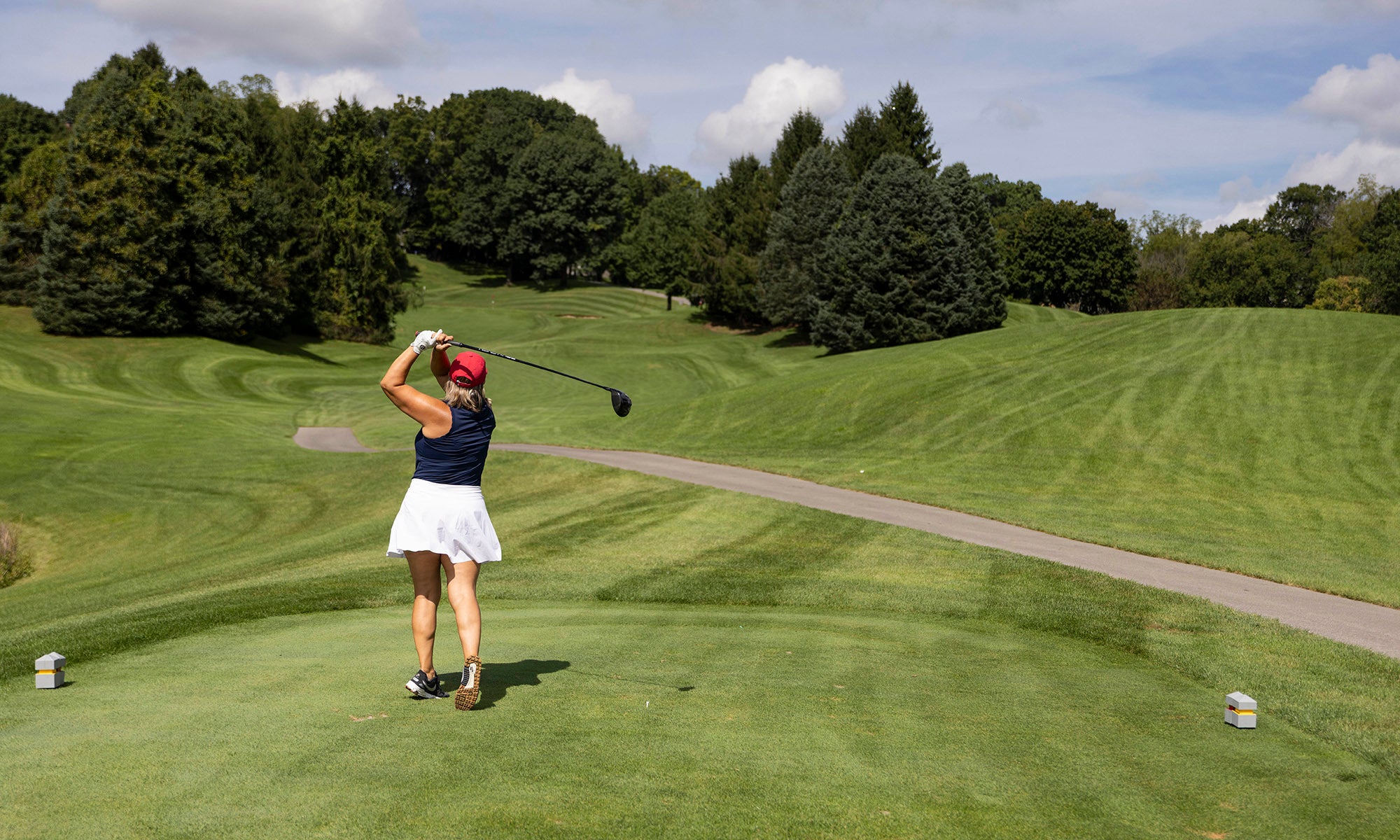 A person wearing a pink cap and white skirt swings a golf club on a green golf course under a partly cloudy sky.