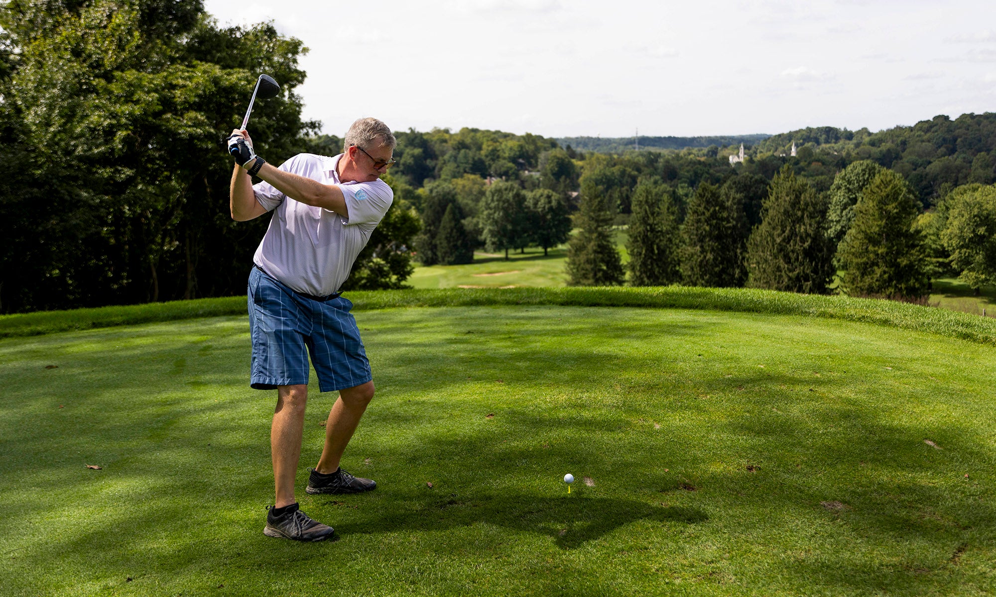 A man wearing a white shirt and blue shorts prepares to swing a golf club on a grassy golf course surrounded by trees.