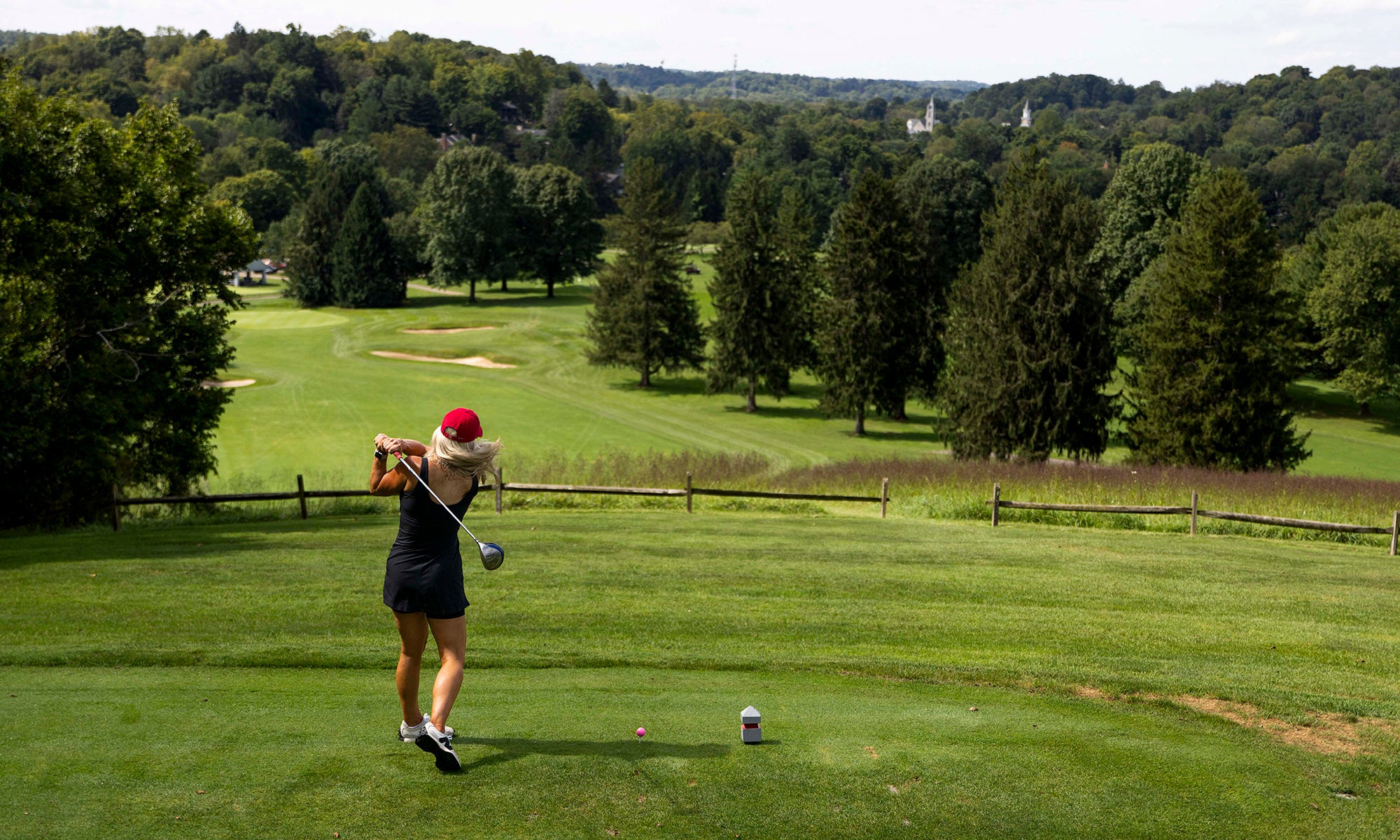 A golfer wearing a red cap swings a club on a green golf course surrounded by trees under a partly cloudy sky.