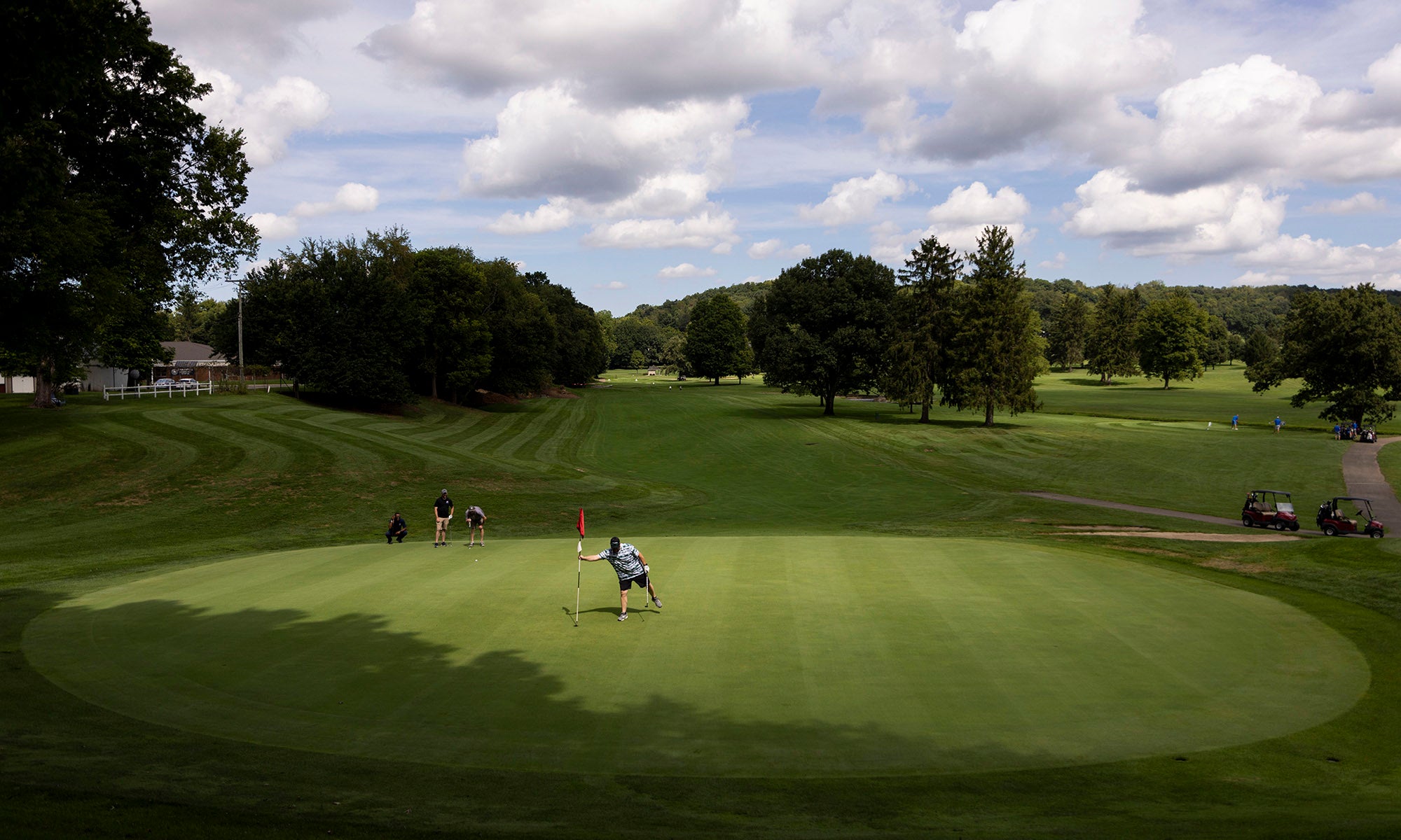 Golfers play on a green surrounded by trees and a rolling golf course under a partly cloudy sky.