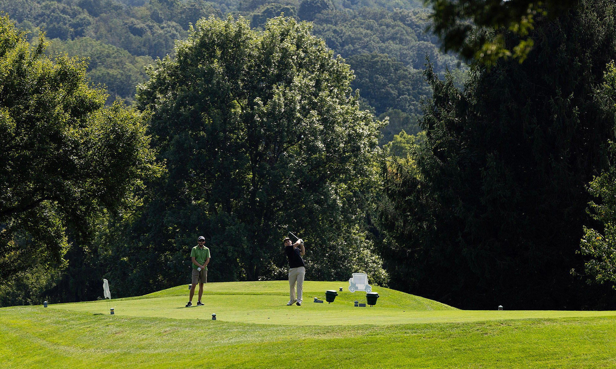 Two people stand on a golf course green surrounded by trees, with golf bags and clubs nearby and wooded hills in the background.