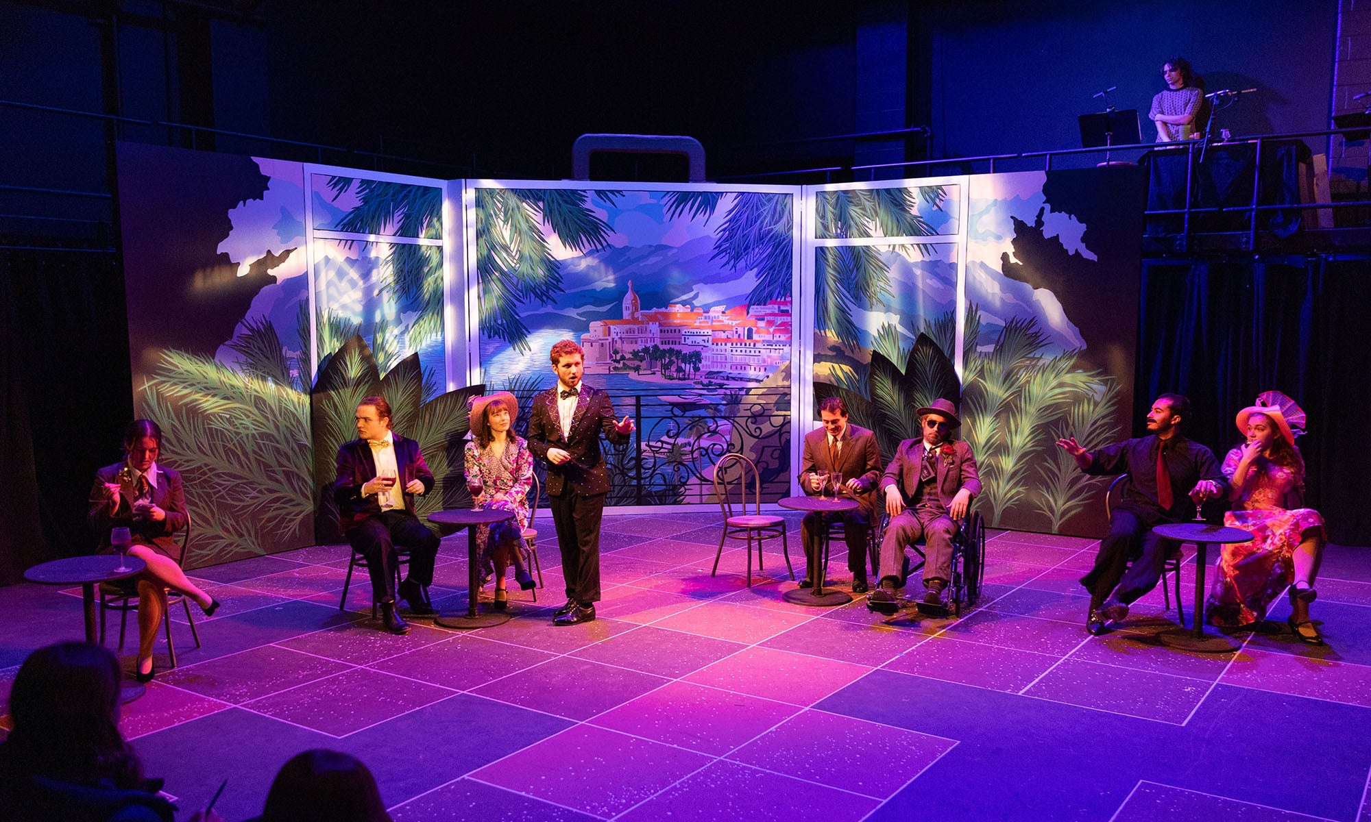Seven actors in formal attire seated and standing on a stage with a painted tropical backdrop, while a person operates equipment on an elevated platform in the background.