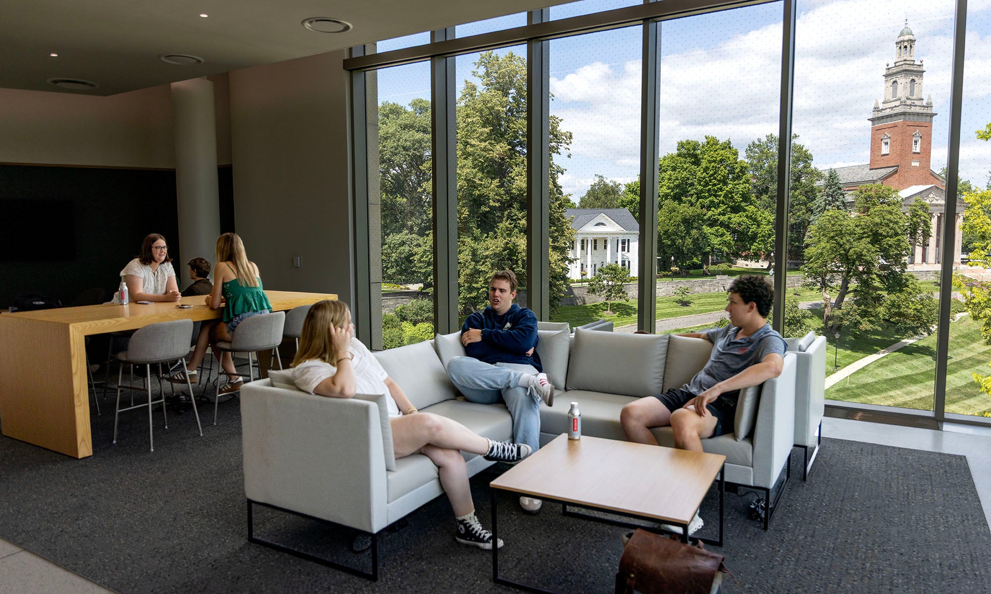 Three people sit on sofas talking in a lounge with large windows overlooking a campus scene; two others work at a high table in the background.