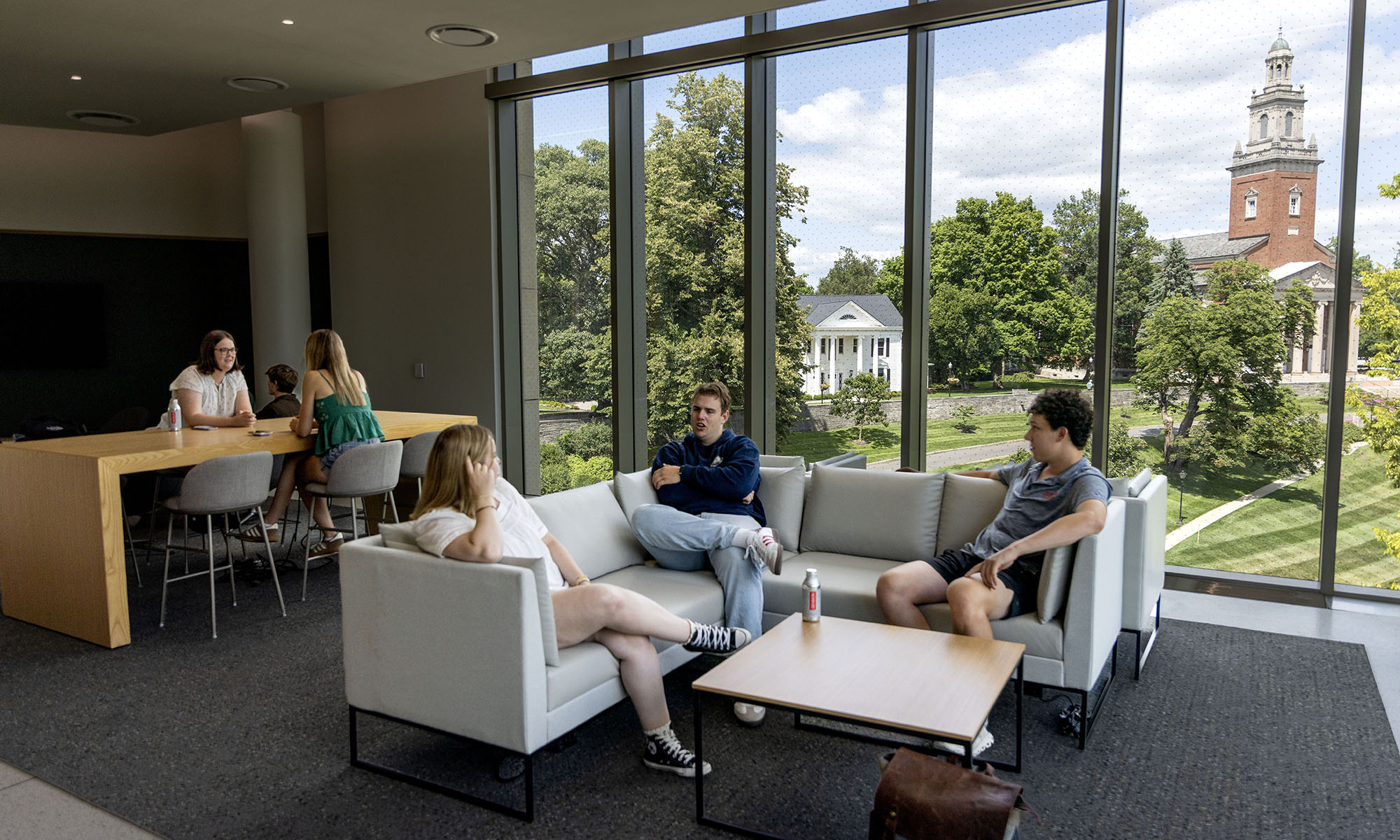 Several people sit and talk in a modern lounge with large windows overlooking a green campus and a tall brick clock tower outside.