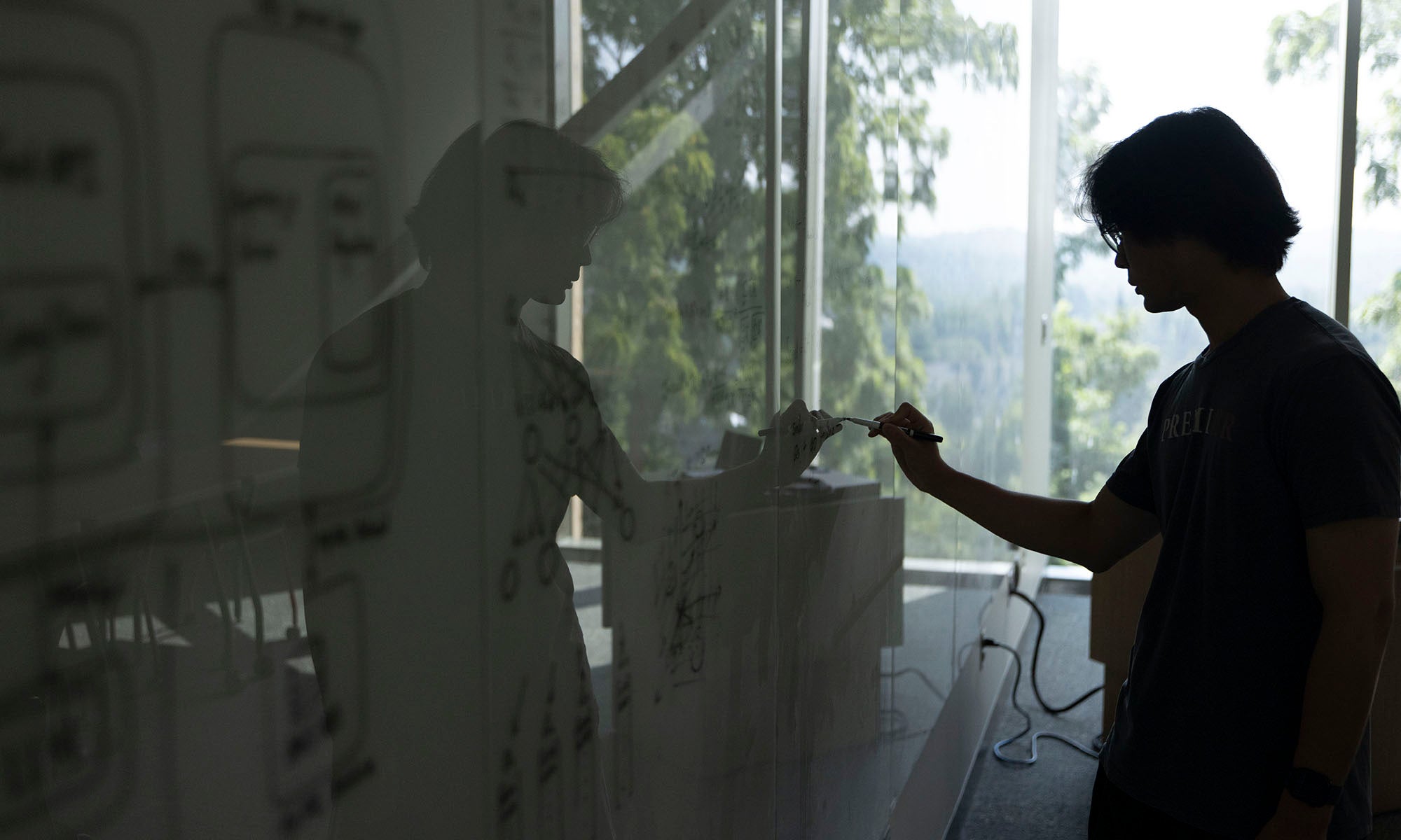 Person writing diagrams or notes with a marker on a glass board in an office with large windows and natural light.