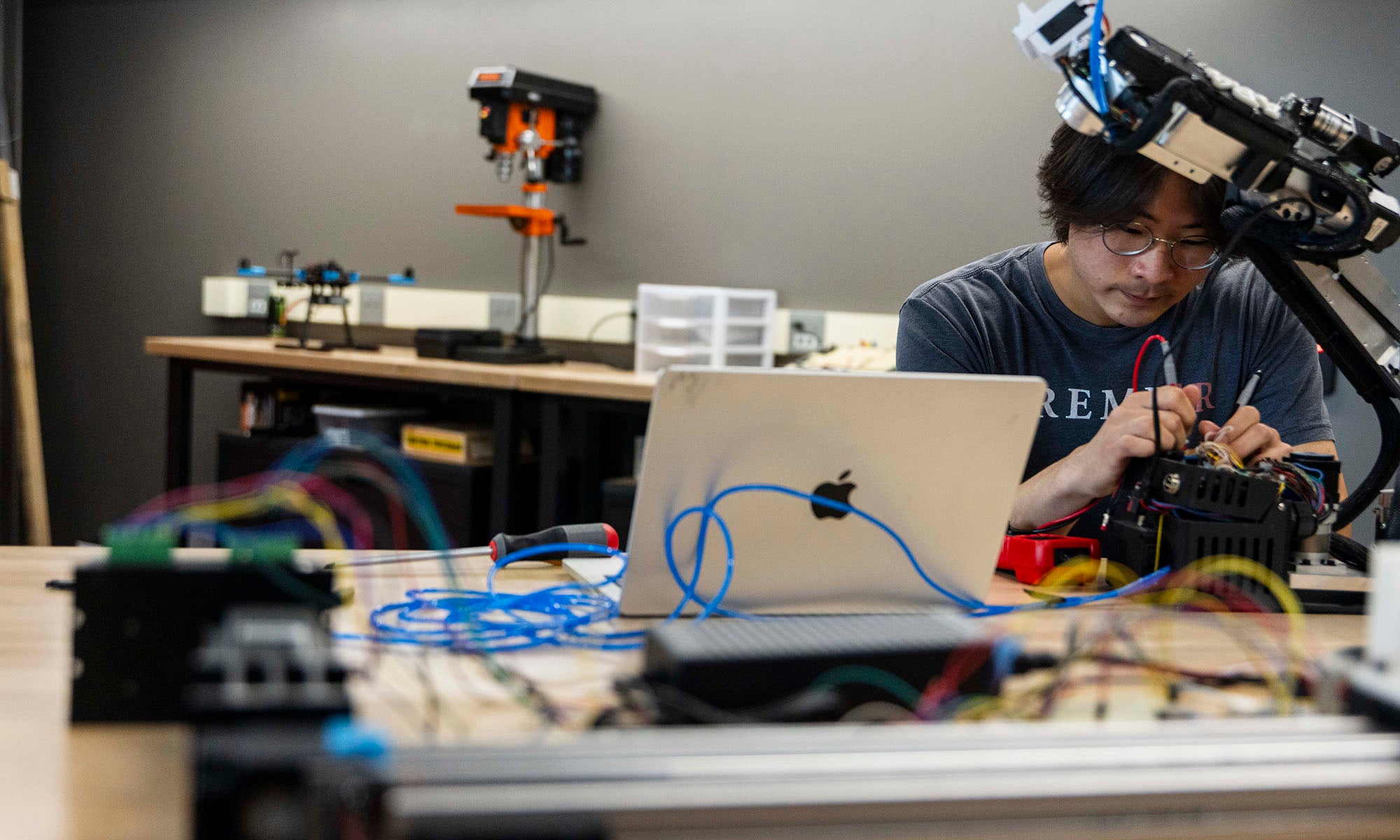 A person works on electronic components at a desk with a laptop and various tools; a robotic arm and other equipment are visible in the workspace.