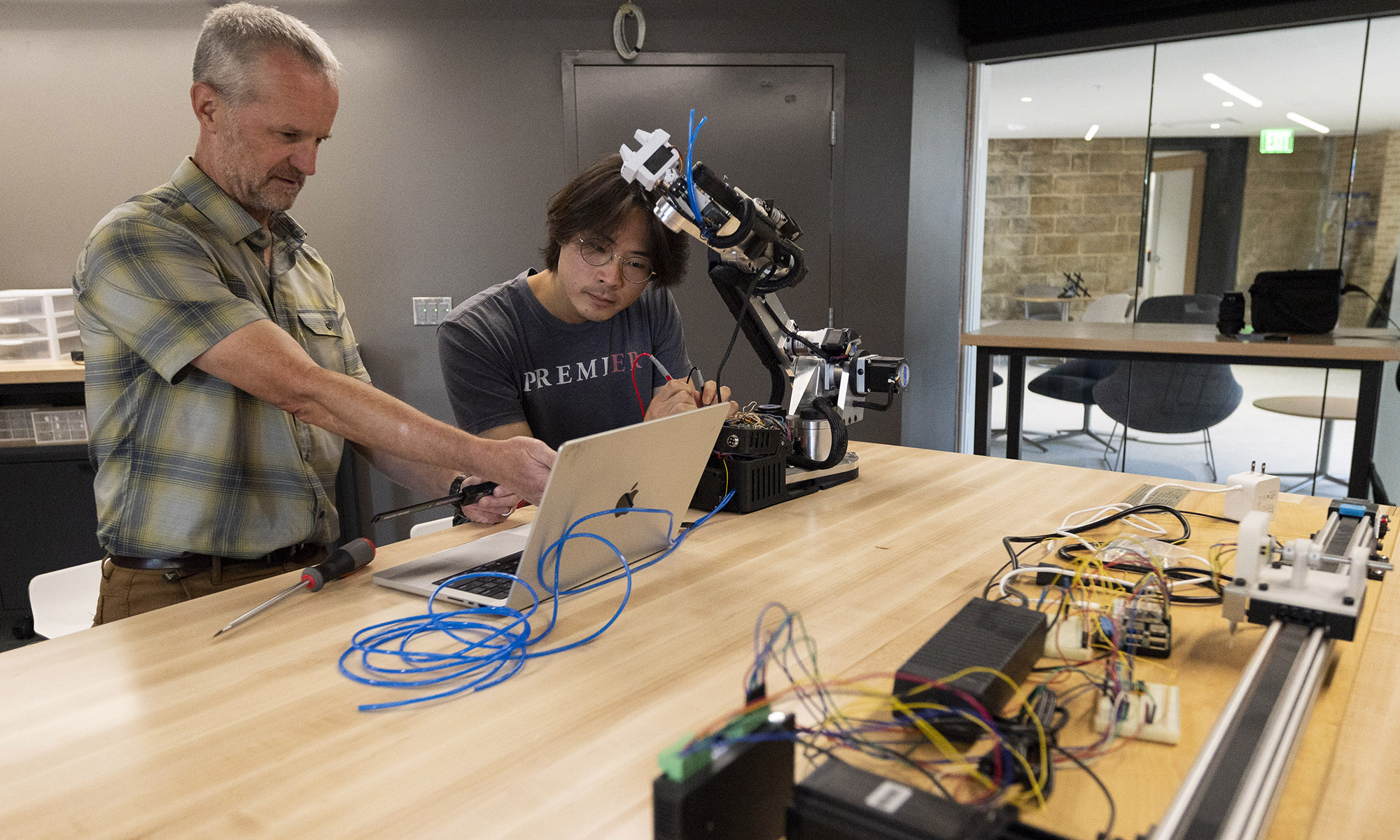 Two people work at a table with a robotic arm, a laptop, and electronic components in a modern lab setting.