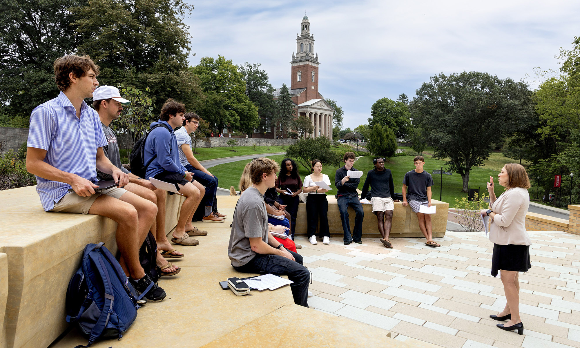 A group of students sits outdoors on stone steps, listening to a woman speaking, with trees and a large building in the background.