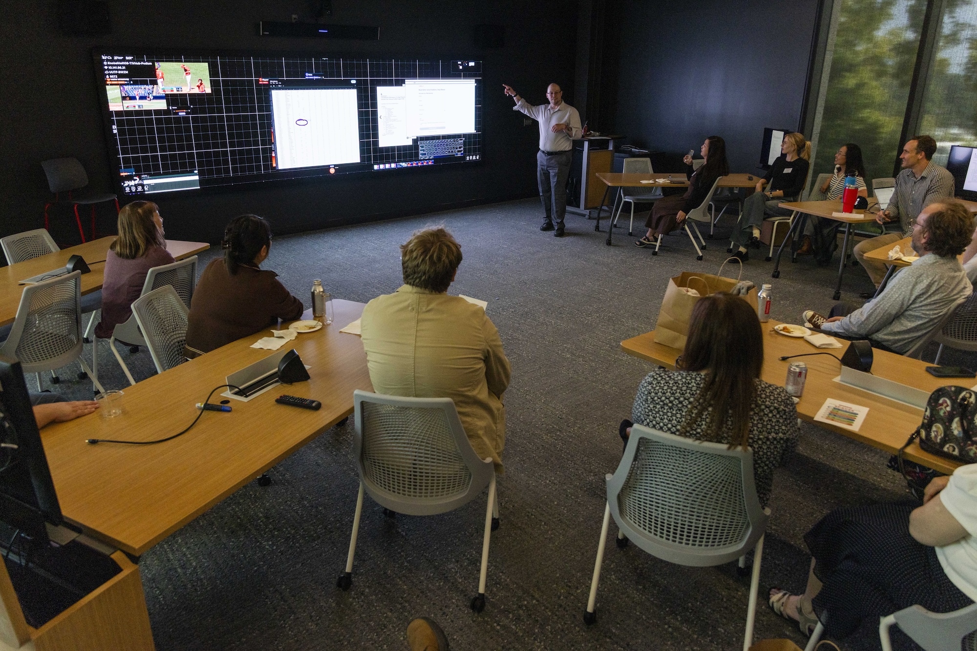 A group of people seated at tables listens to a presenter gesturing toward large screens displaying data and charts