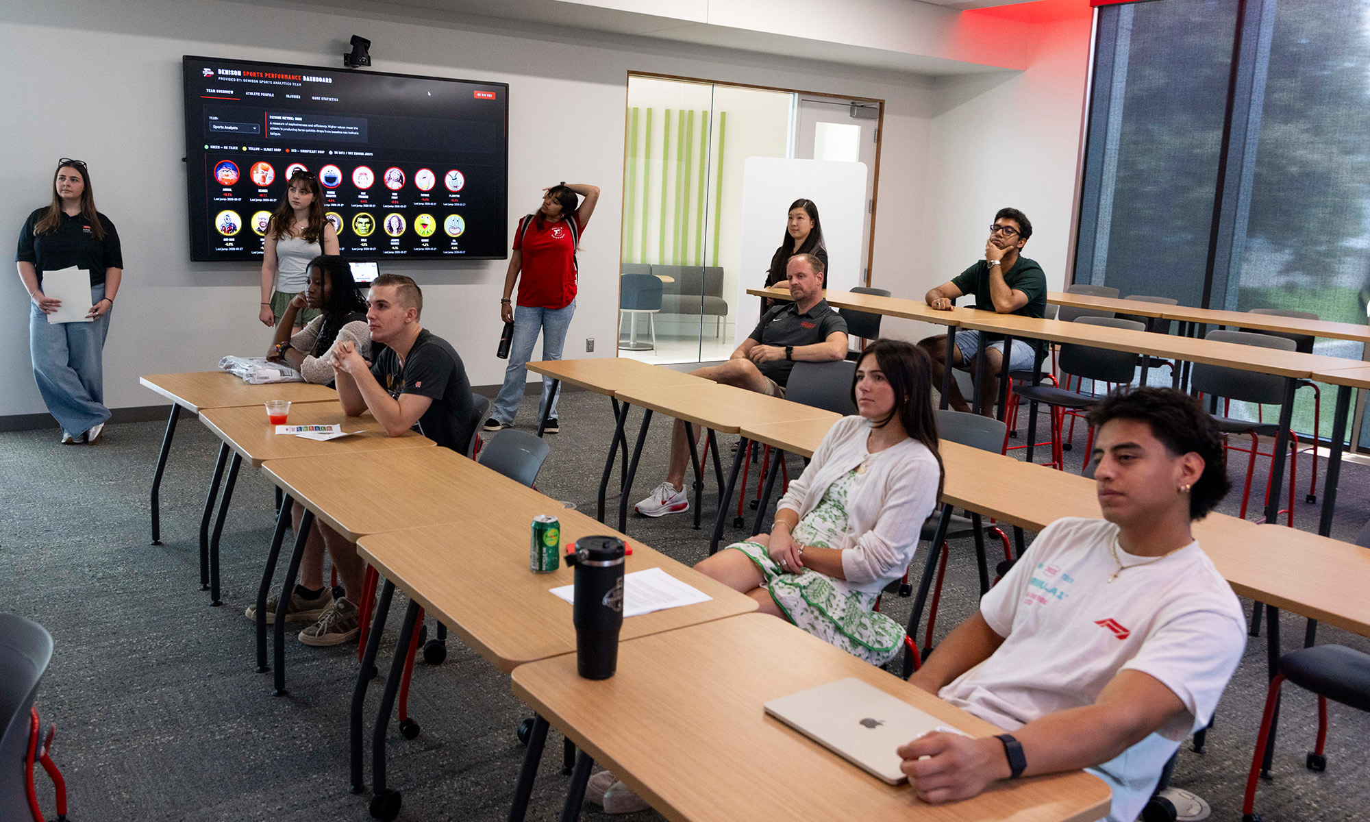 Students sit at desks in a classroom, listening to a presentation displayed on a large screen at the front of the room.