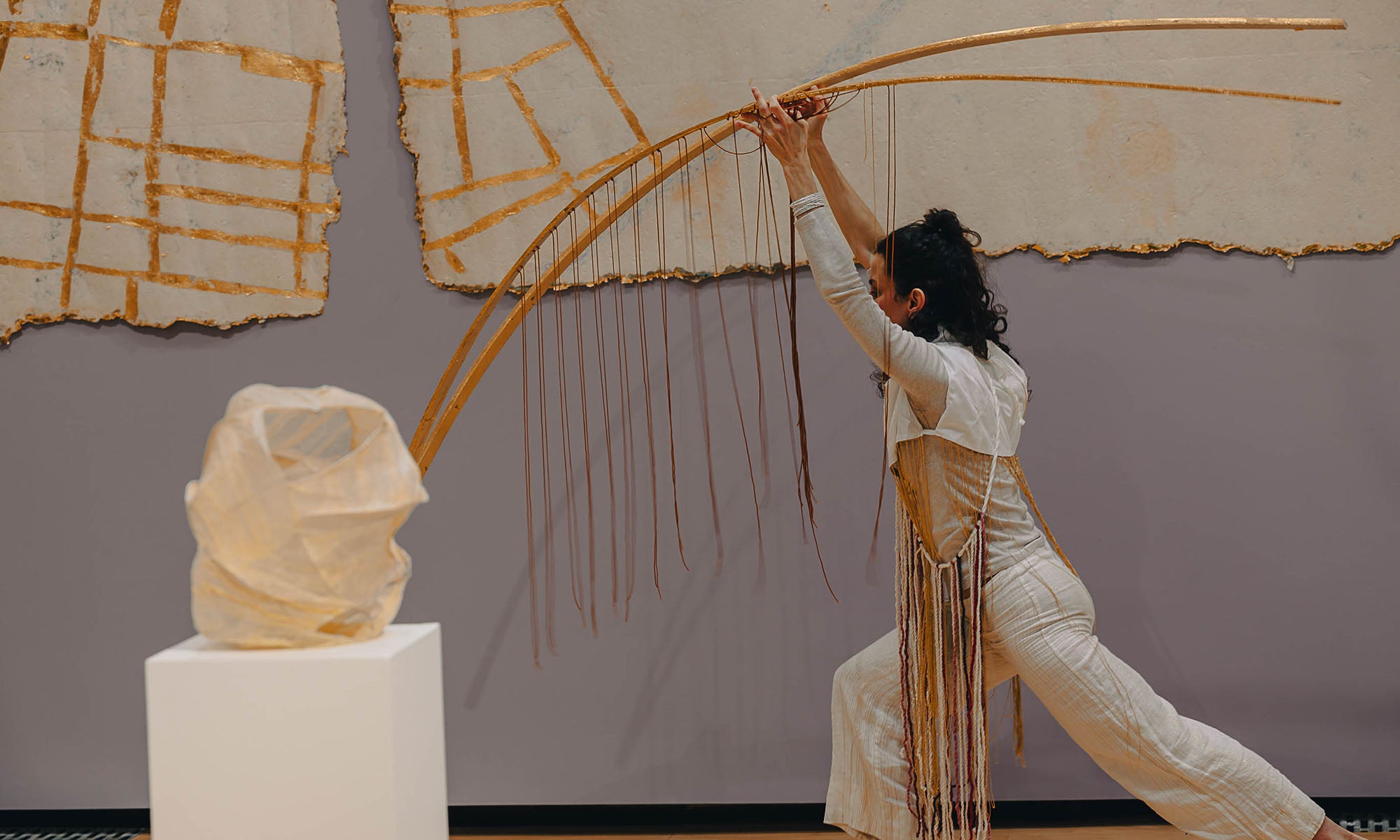 A person in light-colored clothing stretches forward holding two long, curved wooden sticks, in front of abstract art and sculpture on a pedestal, all in colors and textures that match the person's clothing.
