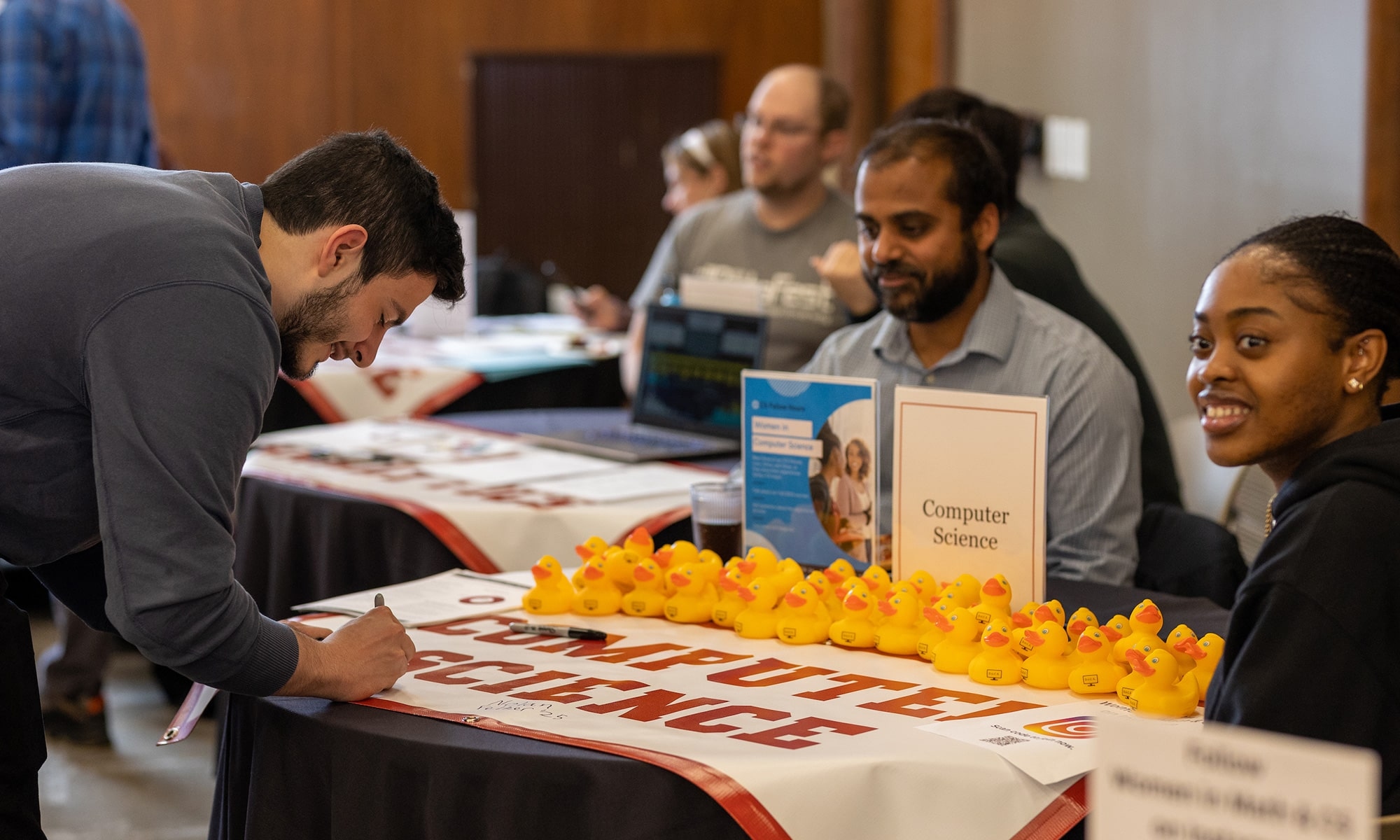 A student signs the Computer Science banner on a table lined with rubber ducks. There is a professor and a student sitting behind the table.