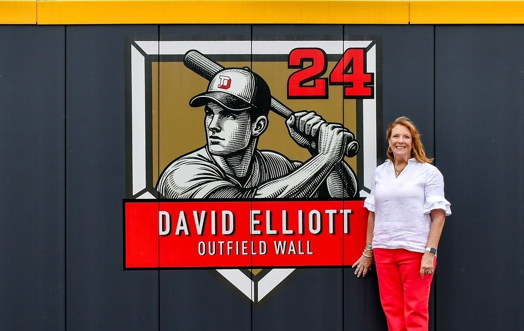 A photo of Lisa Elliott standing by the newly unveiled David Elliott outfield wall graphic.