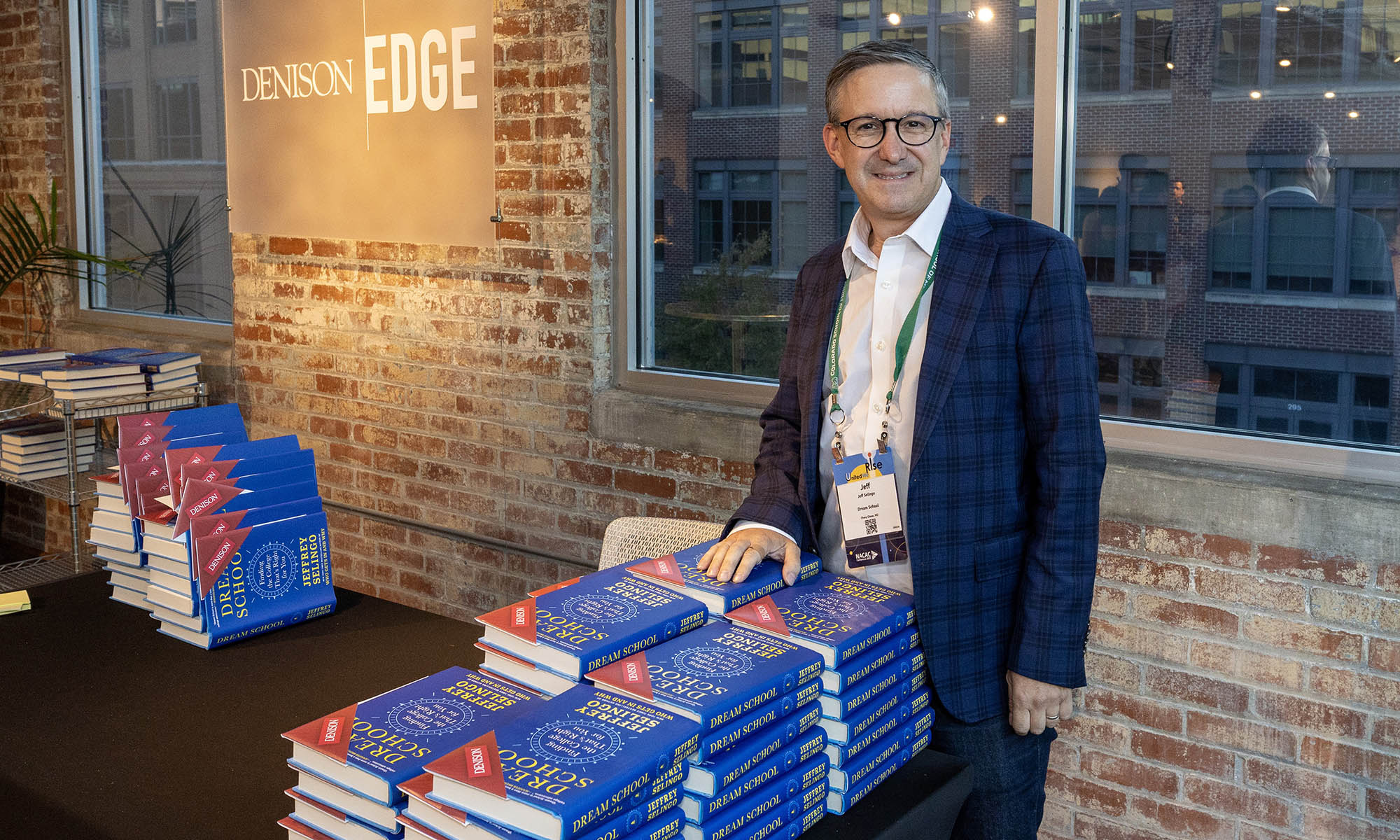 Best-selling author Jeffrey Selingo stands at a table covered with copies of his new book, framed by a brick wall with windows that overlook an urban city center. A sign that reads "Denison Edge" is visible on the brick wall to his right.