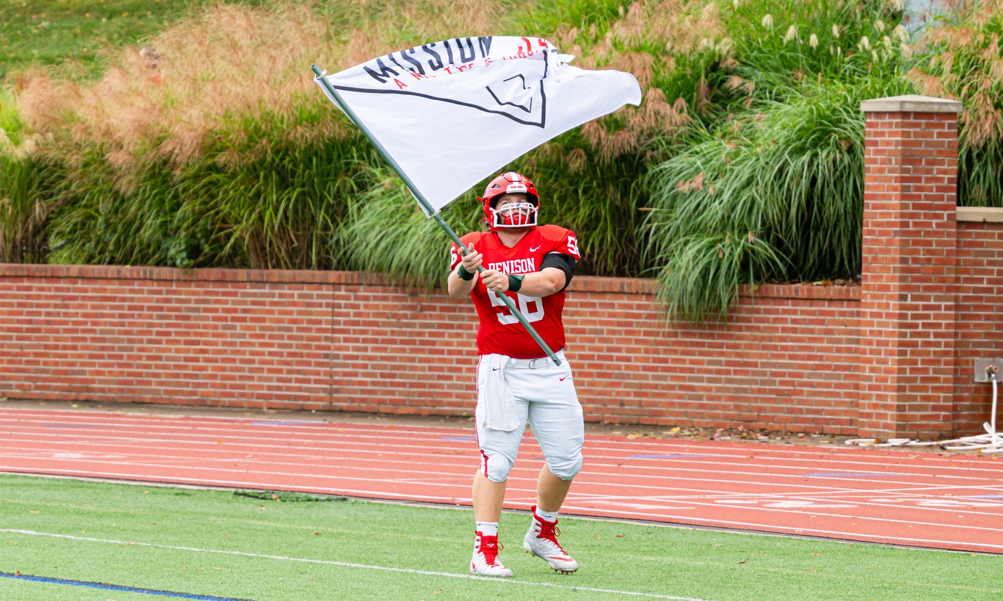 A football player in a red and white uniform stands on a football field waving a large flag bearing the Mission 34 logo.